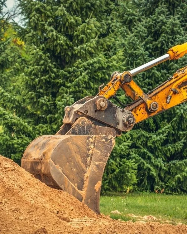 Close-up of a yellow and black excavator bucket digging into the dirt against a background of green trees.