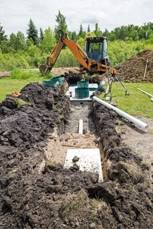 Underground waterline installation site with a small backhoe, pipes, and construction tools in a grassy area surrounded by trees.