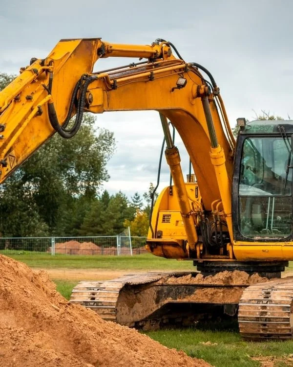 A yellow excavator on a construction site with a mound of dirt in front, greenery, and a chain-link fence in the background.