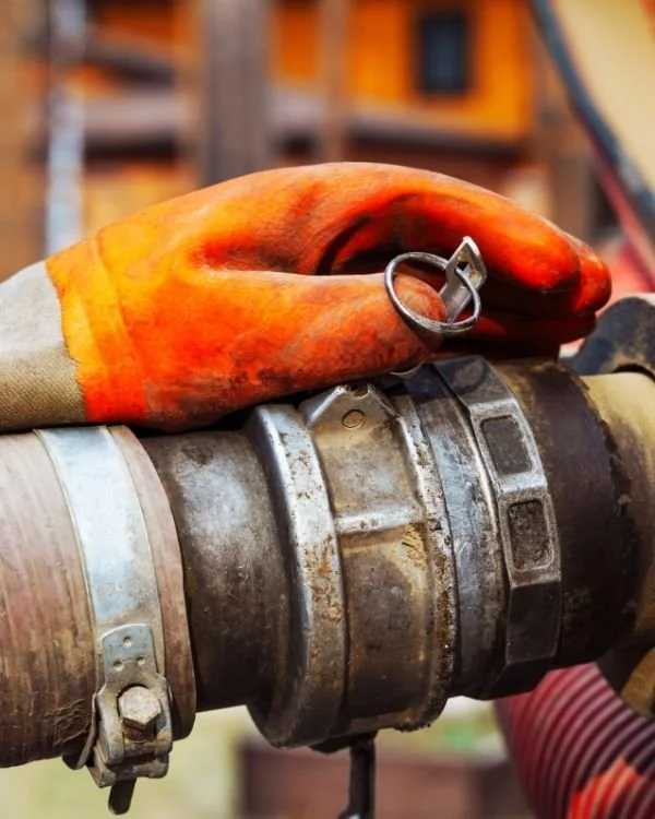 Orange safety glove resting on a metal pipe with a circuit breaker, with a warehouse or workshop in the background.