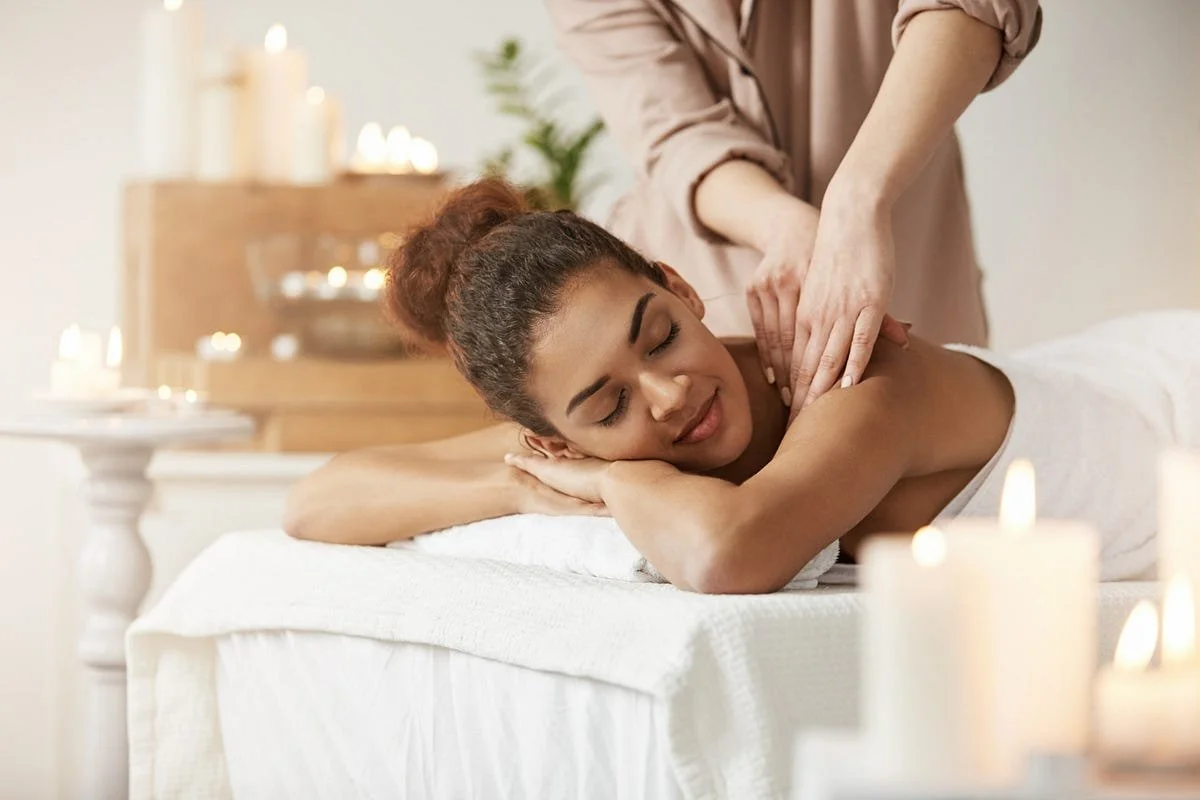 A woman receiving a back massage in a spa or massage therapy setting, lying on her stomach with eyes closed, surrounded by lit candles for relaxation.