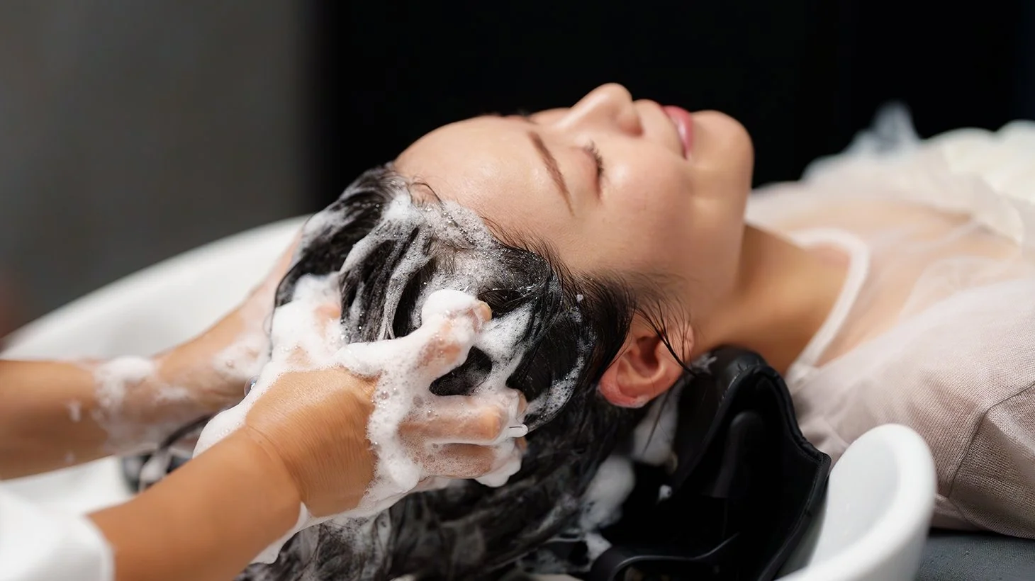 Woman receives a scalp wash at a salon, lying back with her eyes closed, as a stylist shampoos her hair with foam.