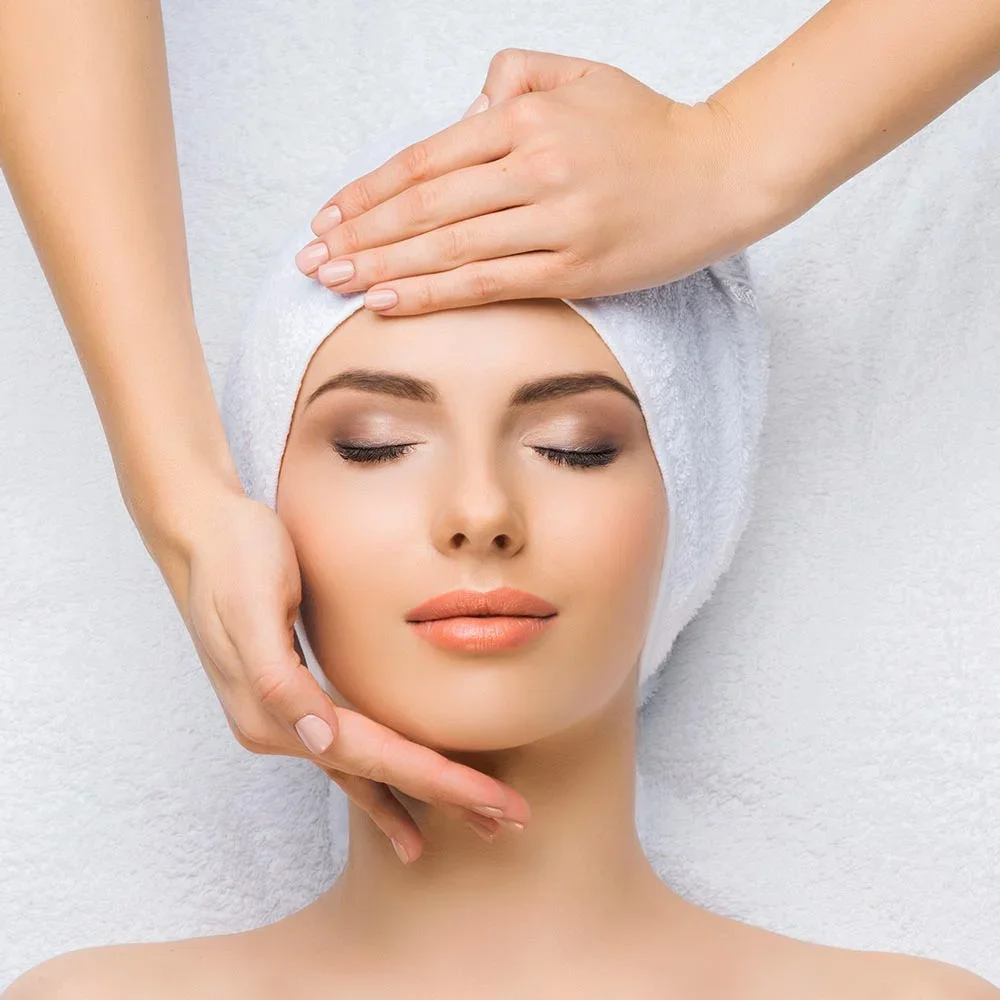 A woman receiving a facial massage at a spa, with her eyes closed and a towel wrapped around her head.