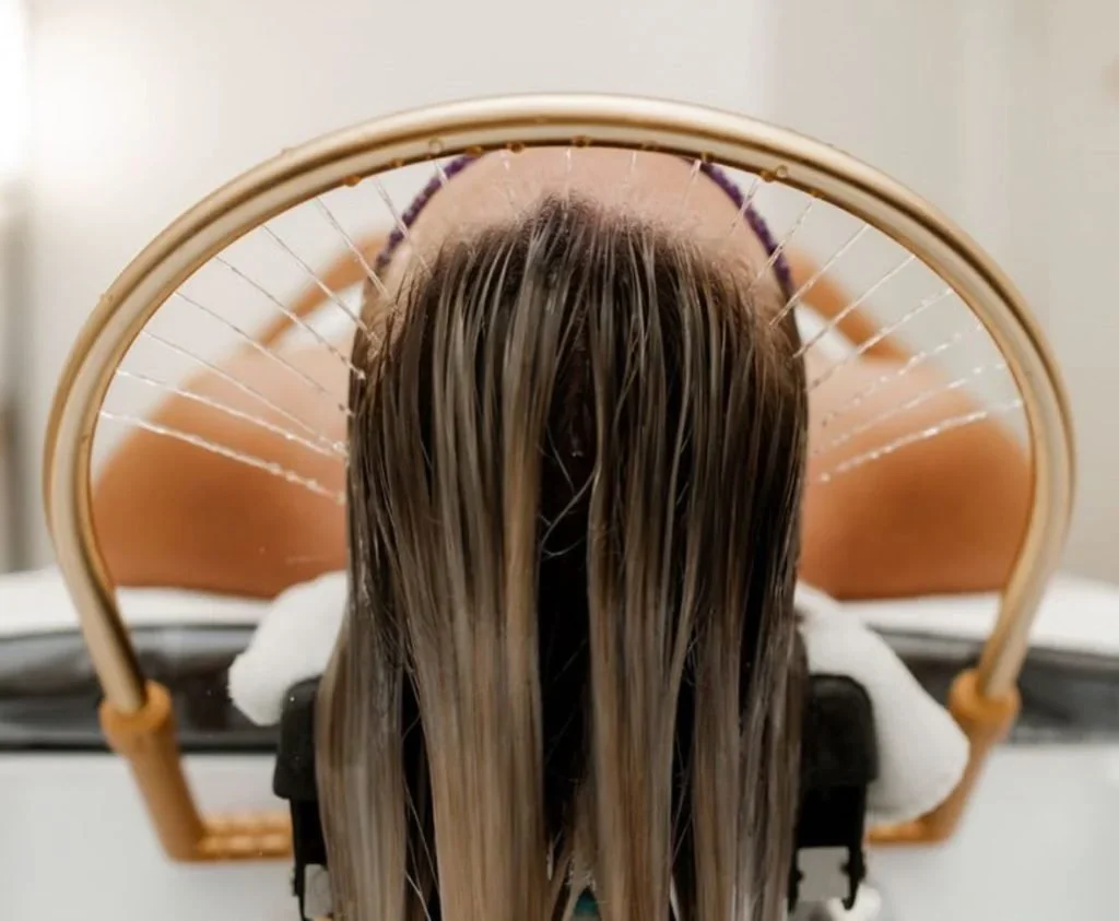 A person receiving a scalp treatment or head spa with water or oil being sprayed onto their hair, viewed from above through a mirror, with the person's head resting on a headrest.