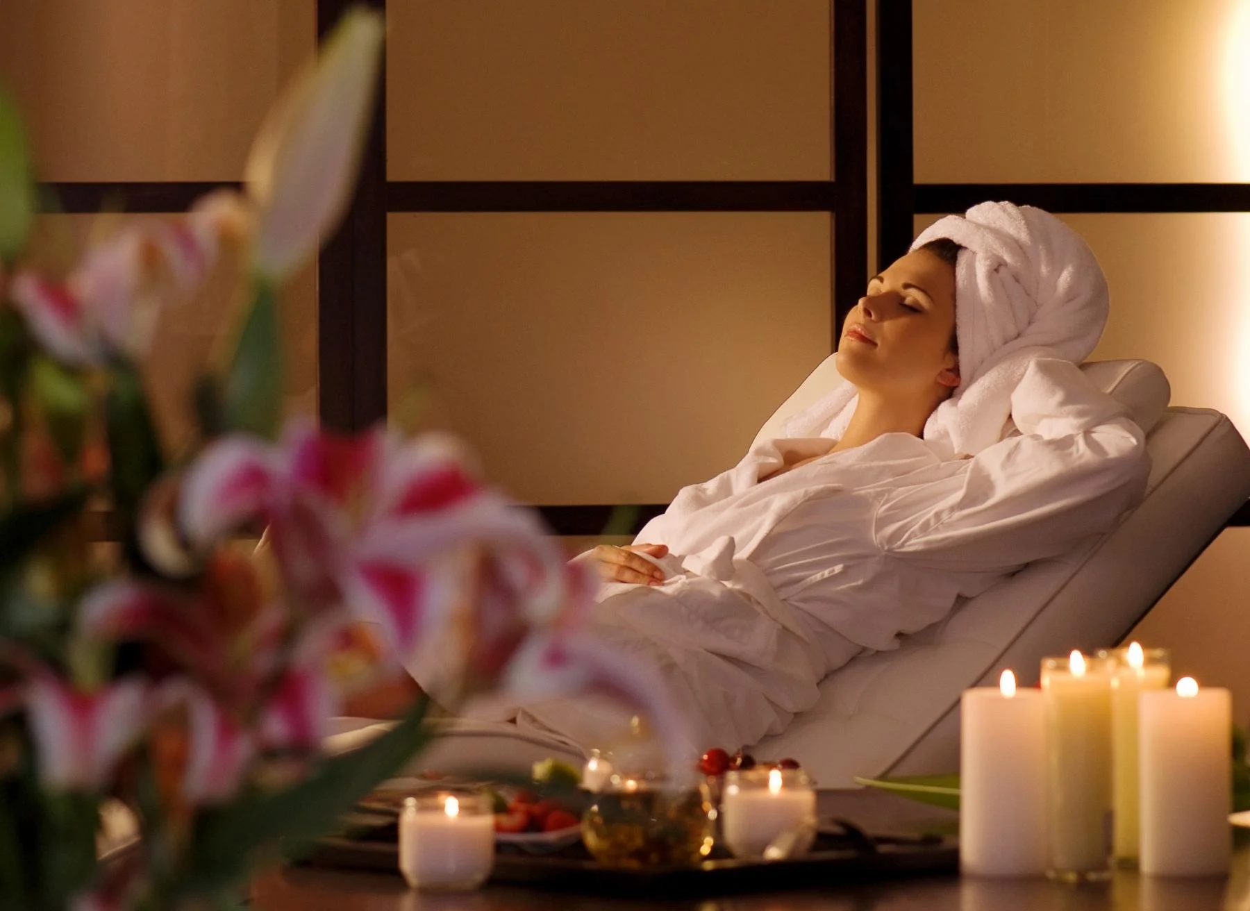 A woman relaxing in a spa or wellness center, lying in a reclining chair with her eyes closed, wrapped in a white robe and with a towel wrapped around her head, surrounded by candles and flowers.