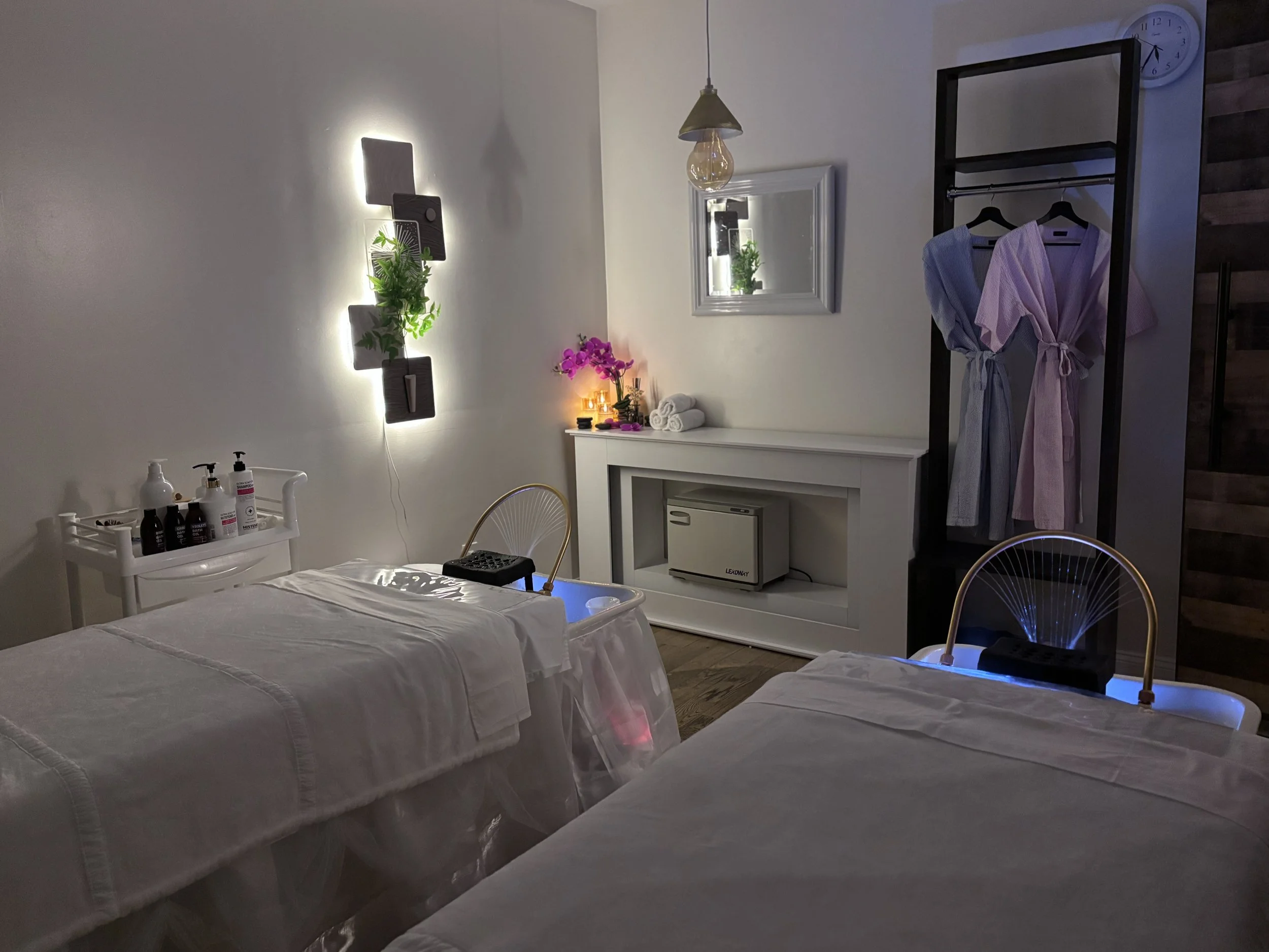 Spa room with two massage tables, a cart with lotion bottles, a wall-mounted plant, a mirror, a small table with candles and flowers, a clothing rack with robes, a clock, and a mini fridge.