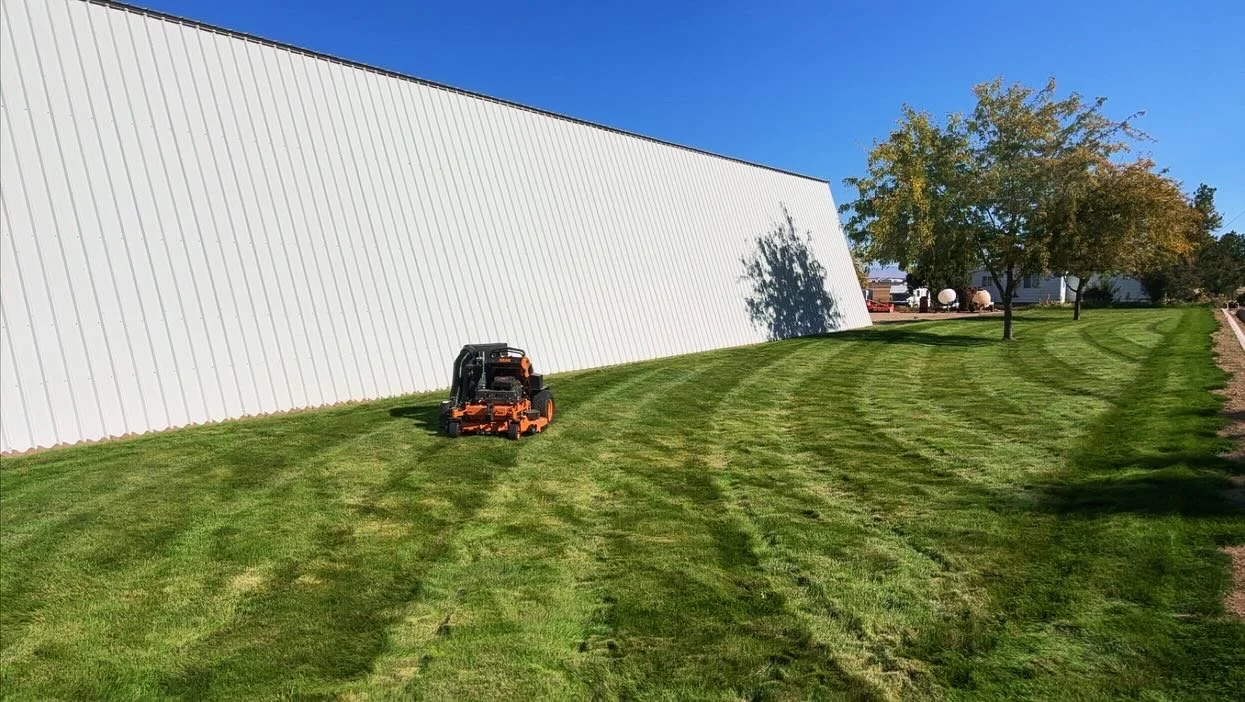 Lawn mower cutting grass on a sloped yard near a white building and trees under a clear blue sky.