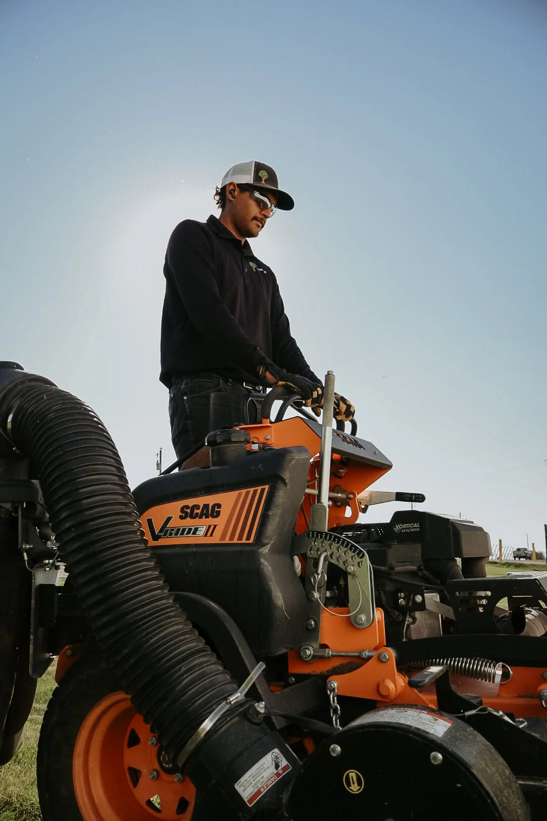A man in sunglasses and a baseball cap operates orange and black agricultural equipment named SCAG V-Ride II outdoors against a clear blue sky.