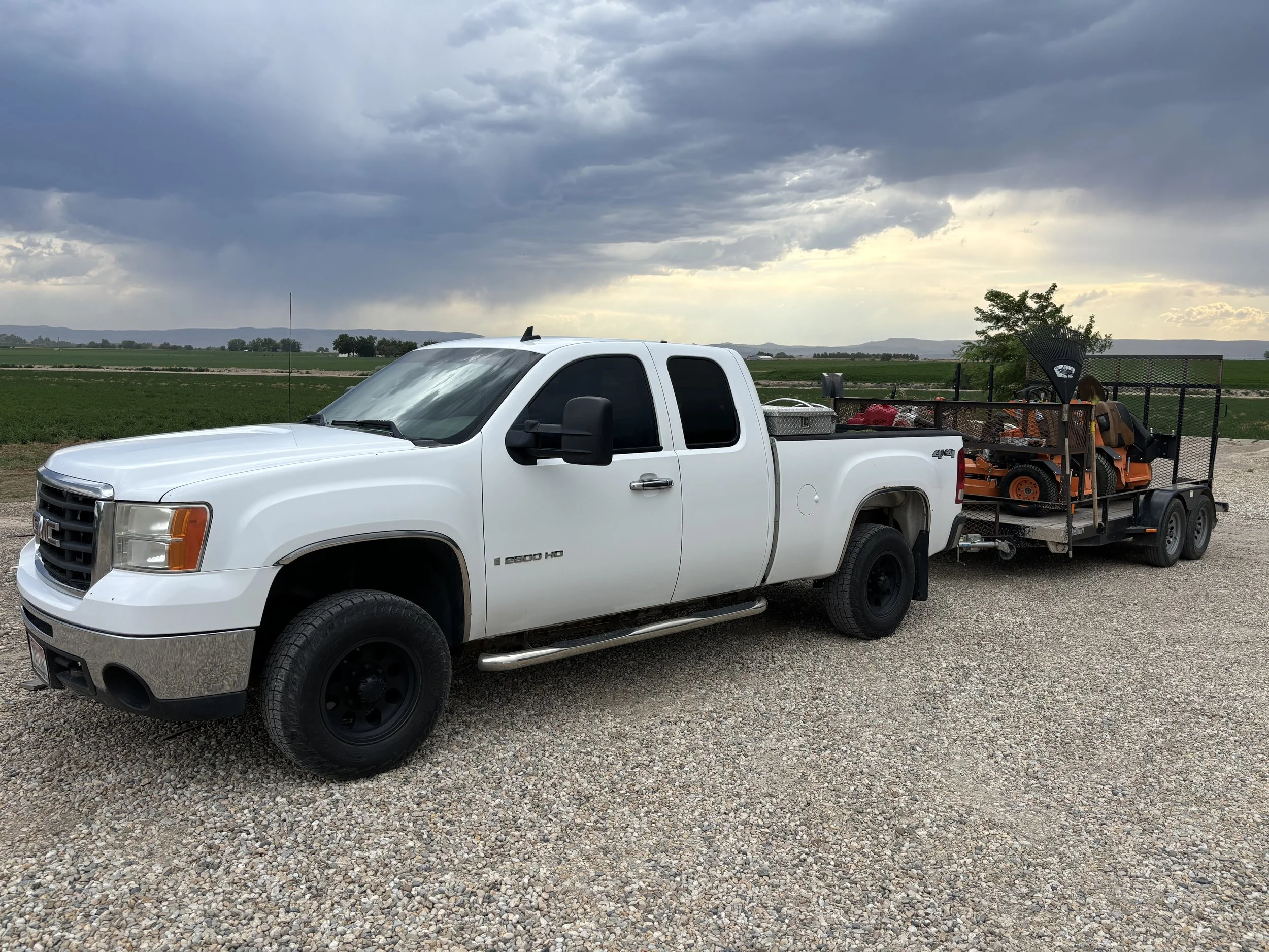 White GMC Sierra pickup truck towing a trailer with outdoor equipment and machinery on it, set against a rural landscape with green fields and cloudy sky.