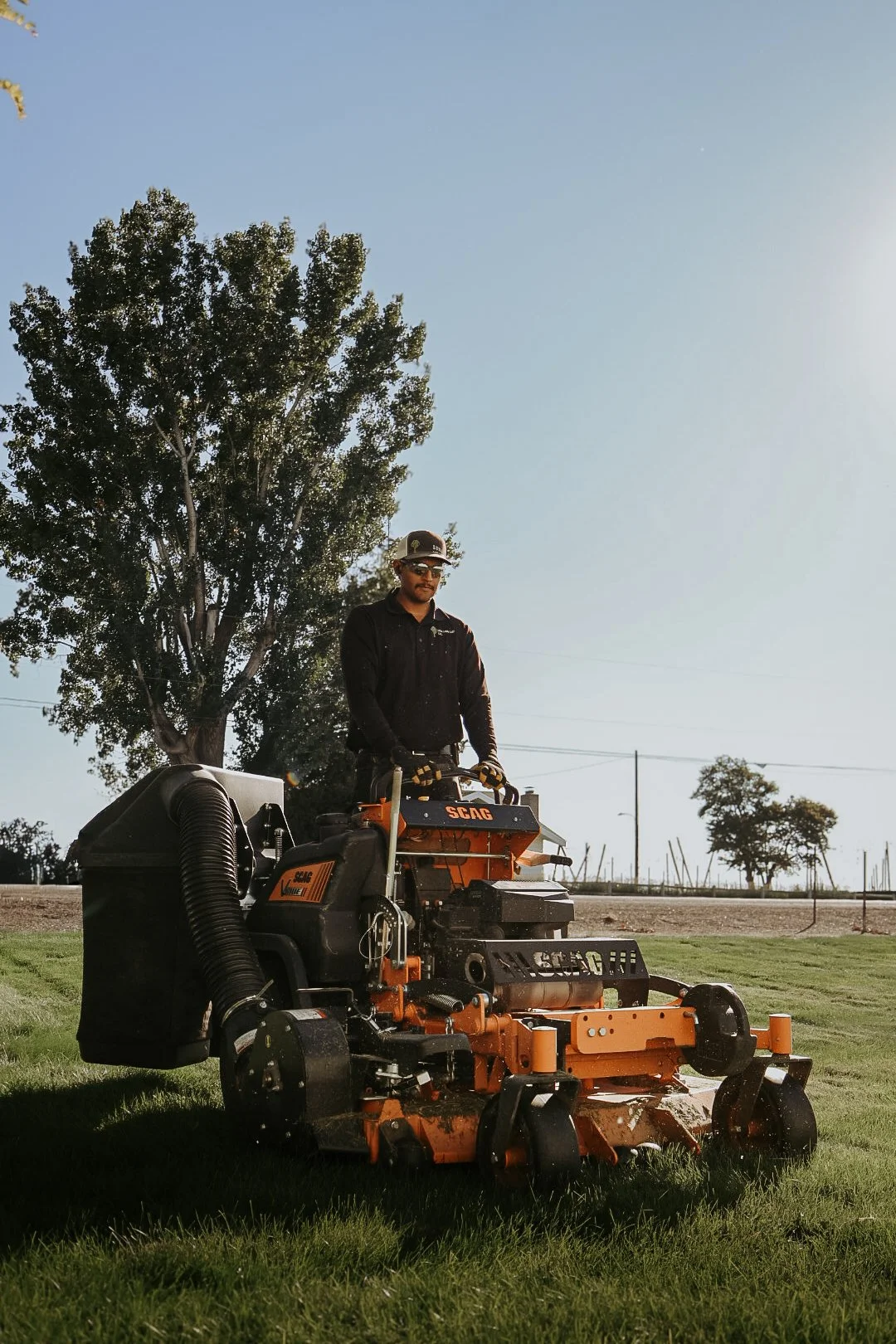 Man operating an orange and black SCAG lawn mower on a grassy field near trees and under a clear blue sky.