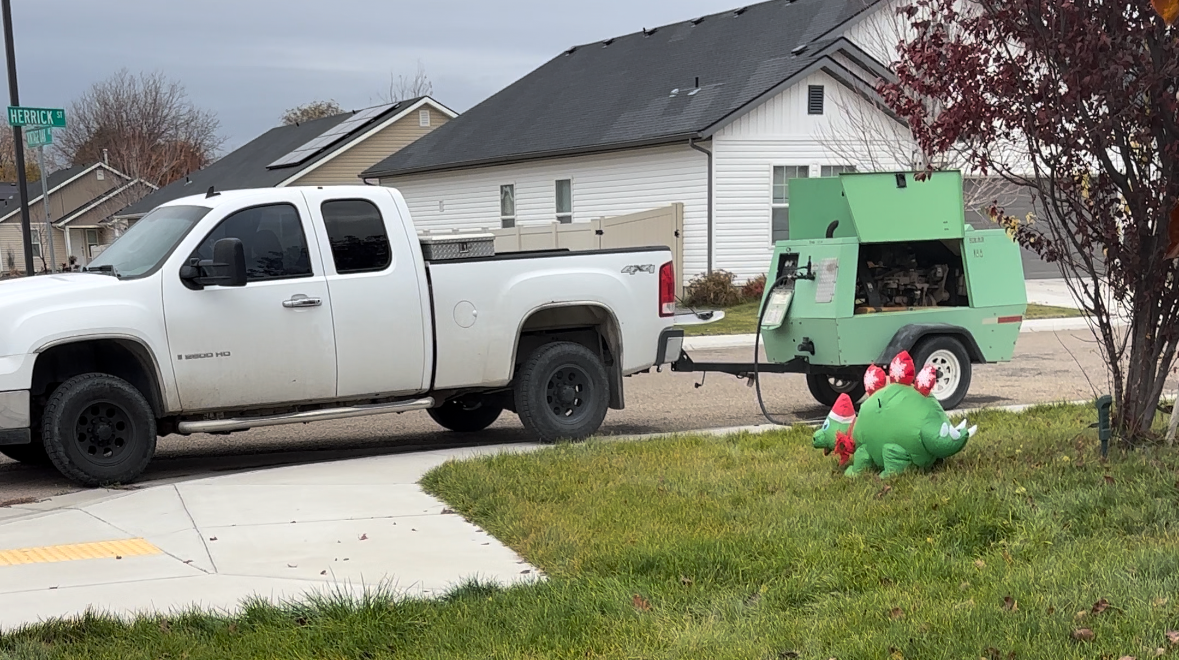 A white pickup truck pulling a green equipment trailer parked on a suburban street, with a decorated inflatable green dinosaur in front of a tree on the lawn.
