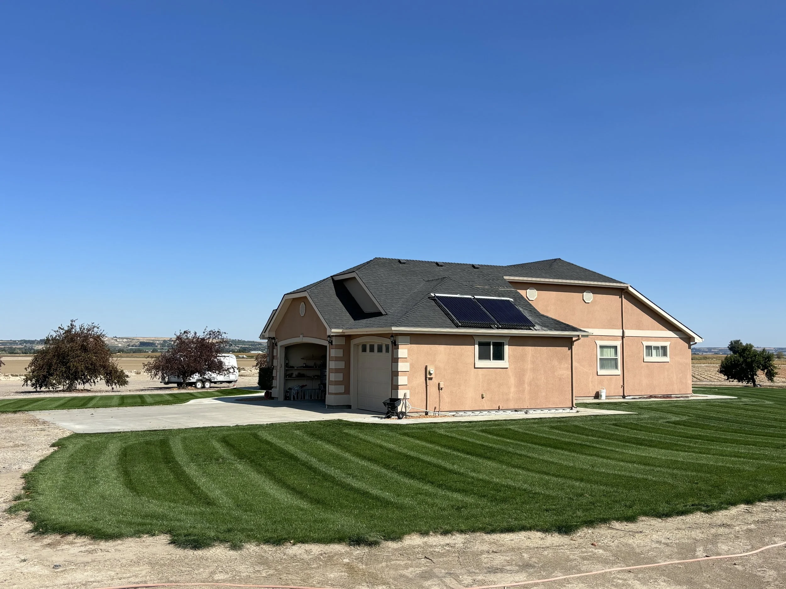 A single-story peach-colored house with a dark gray roof and solar panels, surrounded by lush green grass and a few trees in a rural area under a clear blue sky.