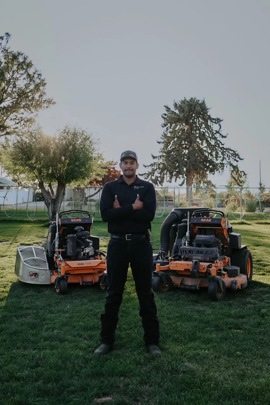 A man in black work attire and a cap standing on a grassy lawn with two large commercial lawn mowers behind him, in a backyard with trees and a fenced area, during sunset.