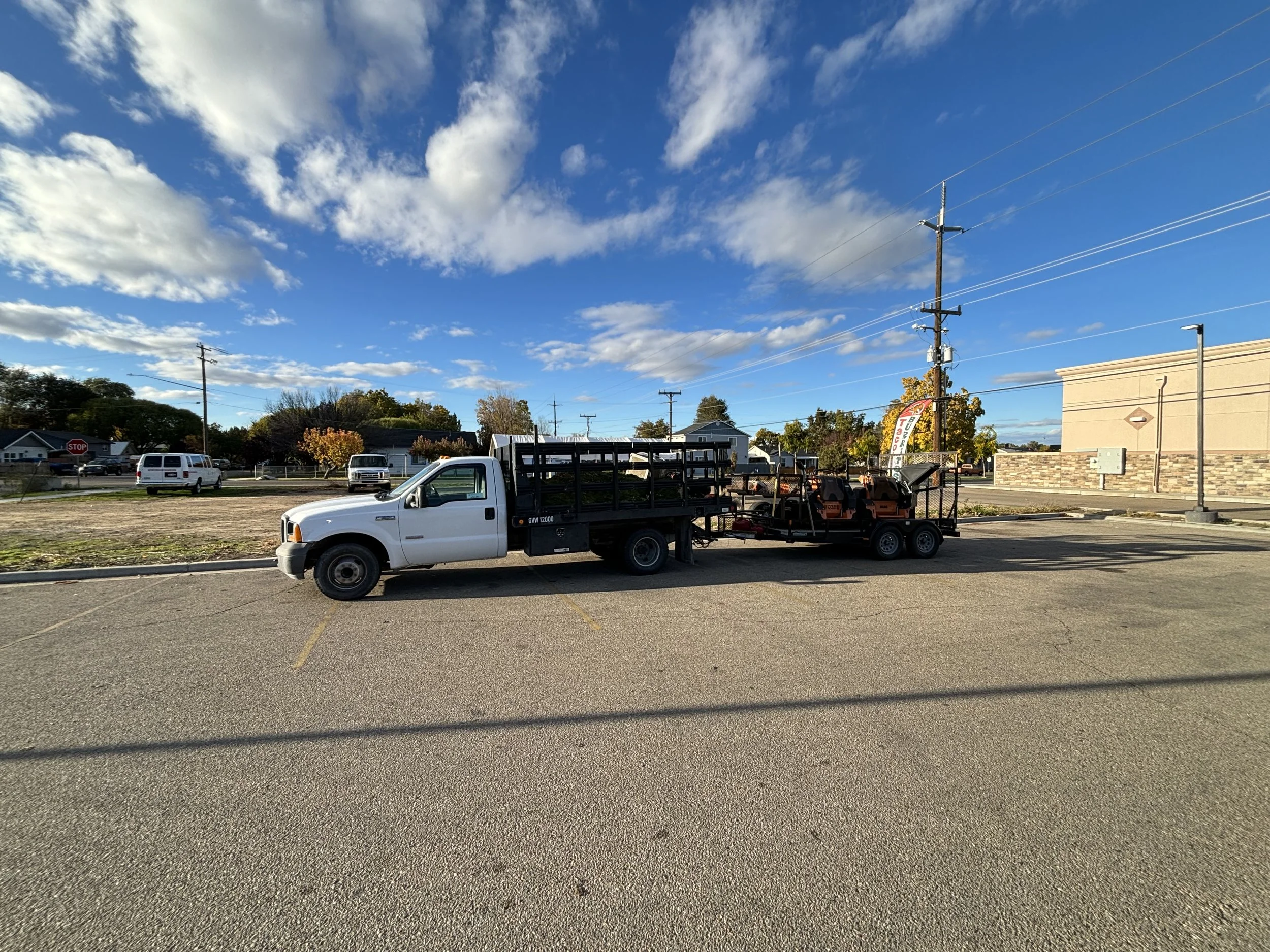 A white pickup truck with a black flatbed trailer carrying equipment parked in an empty parking lot with a clear blue sky and scattered clouds.