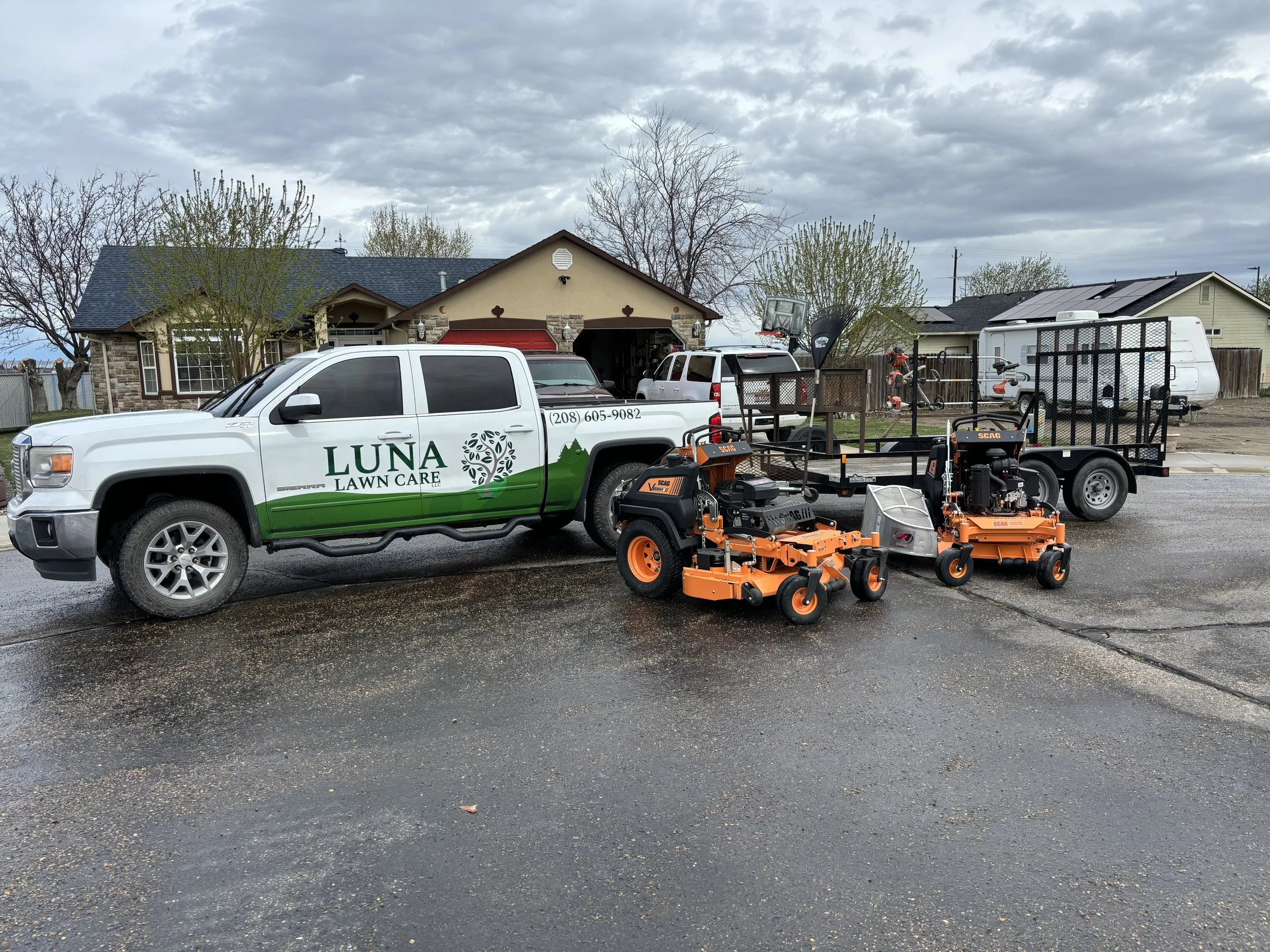 A white pickup truck with green signage reading 'LUNA LAWN CARE' parked on a wet street, with lawn mowing equipment including two orange and black zero-turn mowers, a utility trailer, and other landscaping tools next to it. Residential houses and trees are visible in the background under cloudy skies.