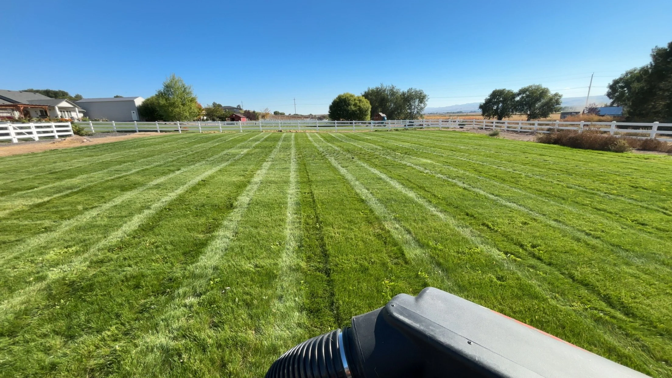 A green grassy field being mowed with a large lawn mower. The sky is clear and blue, with some trees and houses in the background.