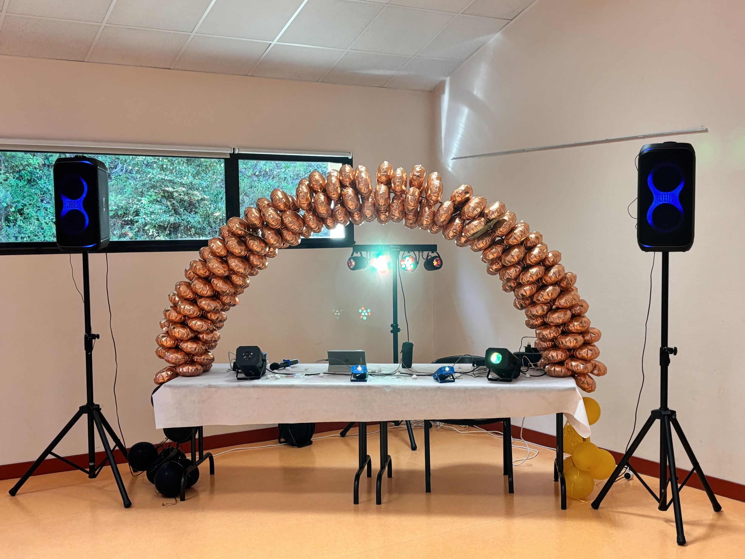 Décor de fête avec une arche en ballons dorés, deux enceintes bleues sur des supports, une table avec des équipements musicaux, et des ballons jaunes au sol.