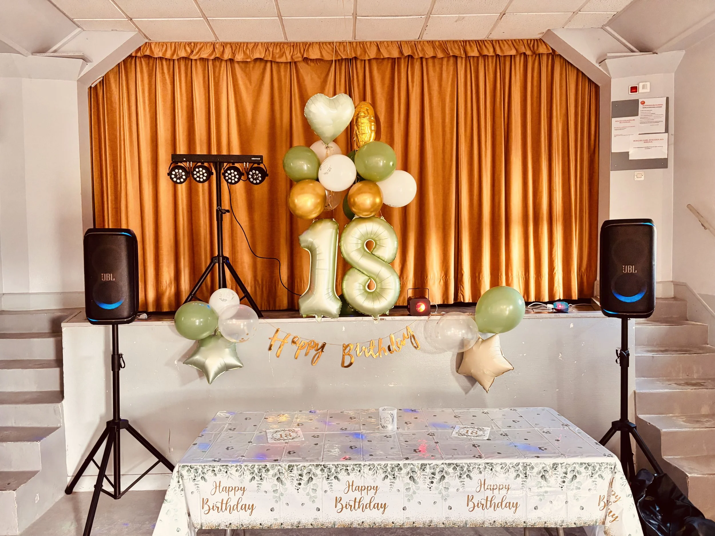 Décor de fête d'anniversaire avec des ballons dorés, verts et blancs, un grand chiffre 18 en ballons et une bannière "Happy Birthday" sur une table recouverte d'une nappe à motifs.