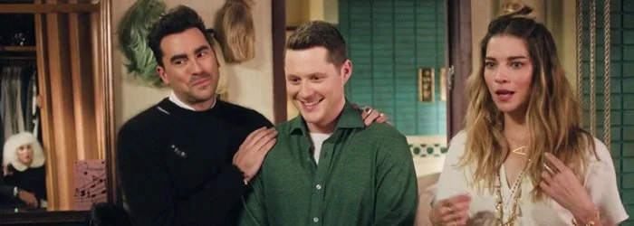 Three people standing together indoors, two men and one woman, with a woman sitting in the background near a piano.