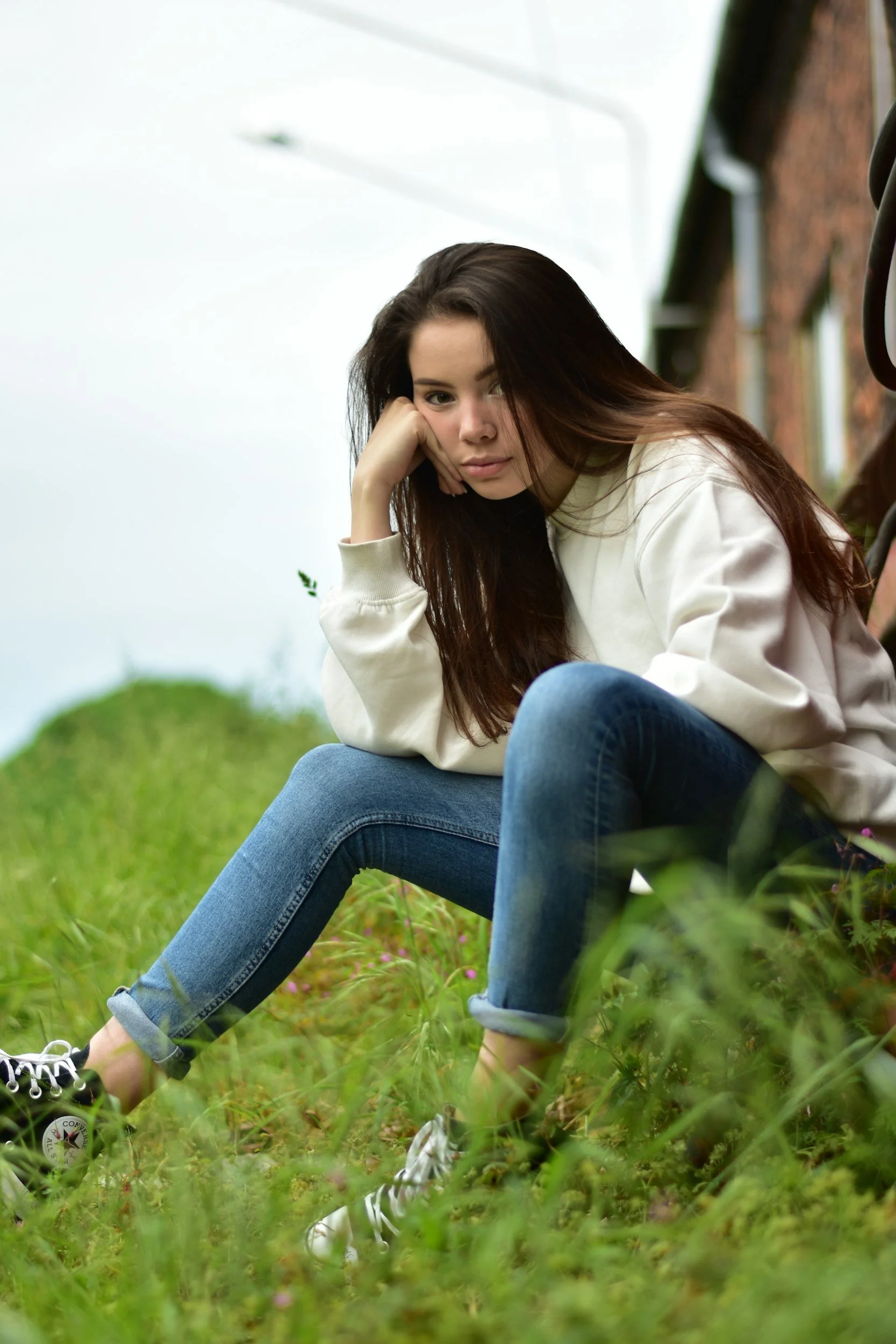 Young woman sitting on grass outdoors near a brick building, wearing a white sweatshirt and blue jeans, with her head resting on her hand, looking at the camera.