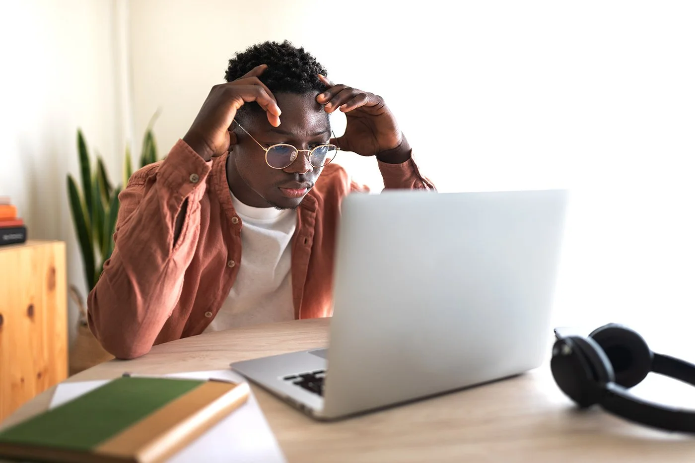 Young man with glasses and curly hair, wearing a brown shirt, looks at a laptop screen with a stressed expression, sitting at a desk with a green and orange notebook, nearby headphones, and a plant in the background.
