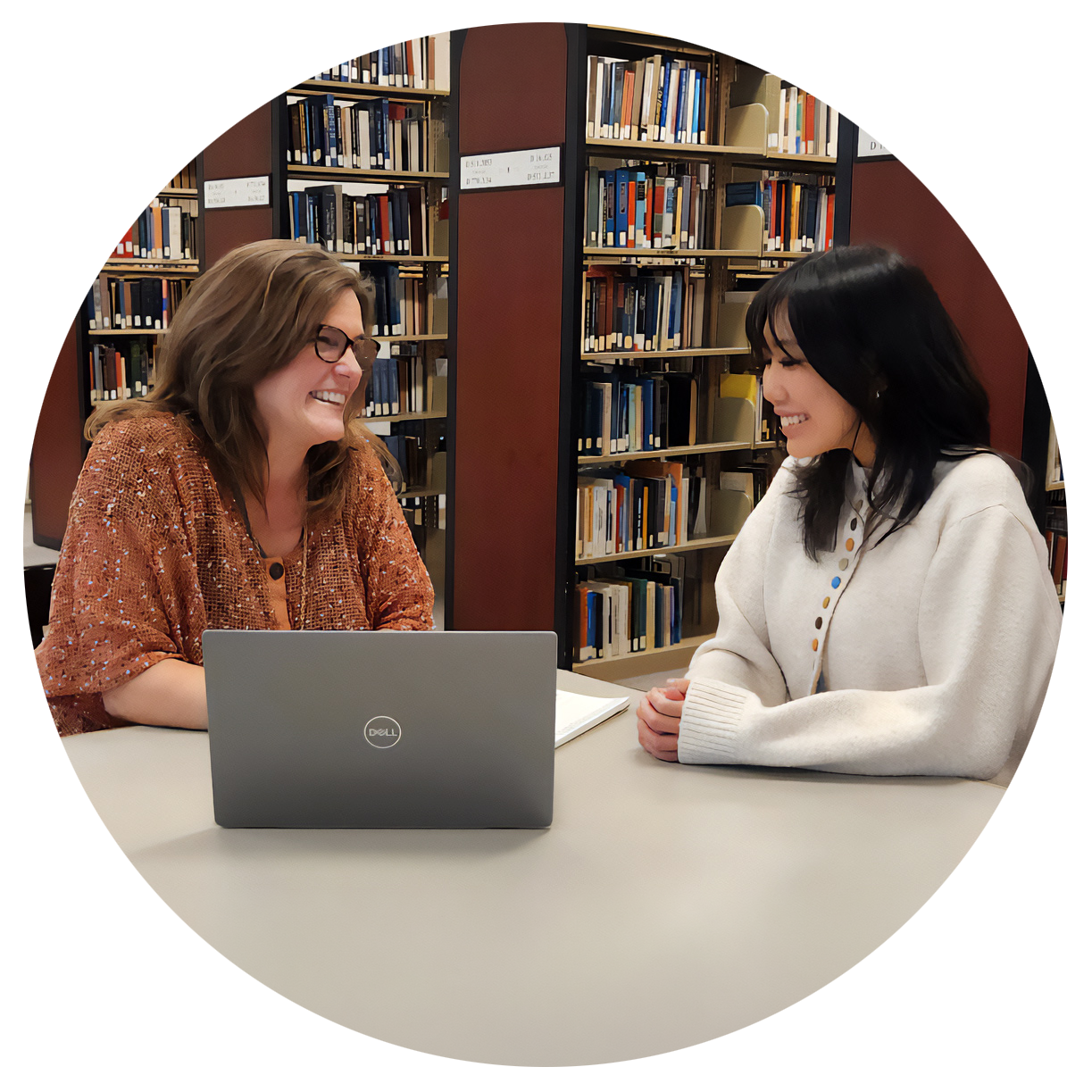 Two women sit at a table in a library, smiling and looking at each other. One woman has brown hair, wears glasses, and a brown sweater. The other woman has black hair and wears a white sweater. There are bookshelves filled with books behind them.