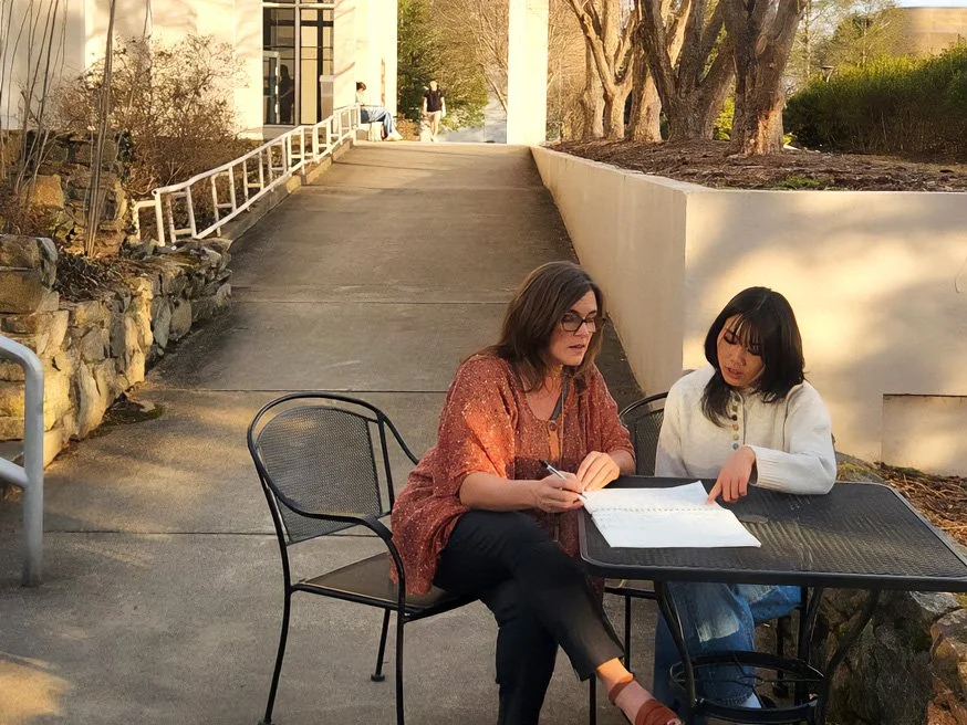 Two women sitting at a black outdoor table, looking at papers, with a white building and trees in the background during daytime.