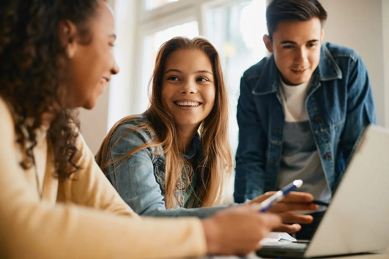 Three teenagers sitting at a table with a laptop, smiling and engaging in conversation.