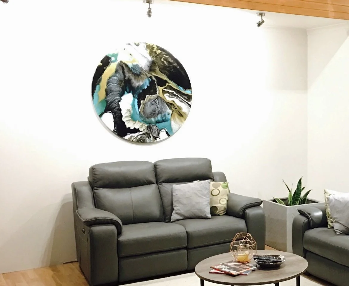 Living room with gray leather sofa, decorative pillows, a small wooden coffee table with candles and magazines, potted plants, abstract wall art, and a white wall.