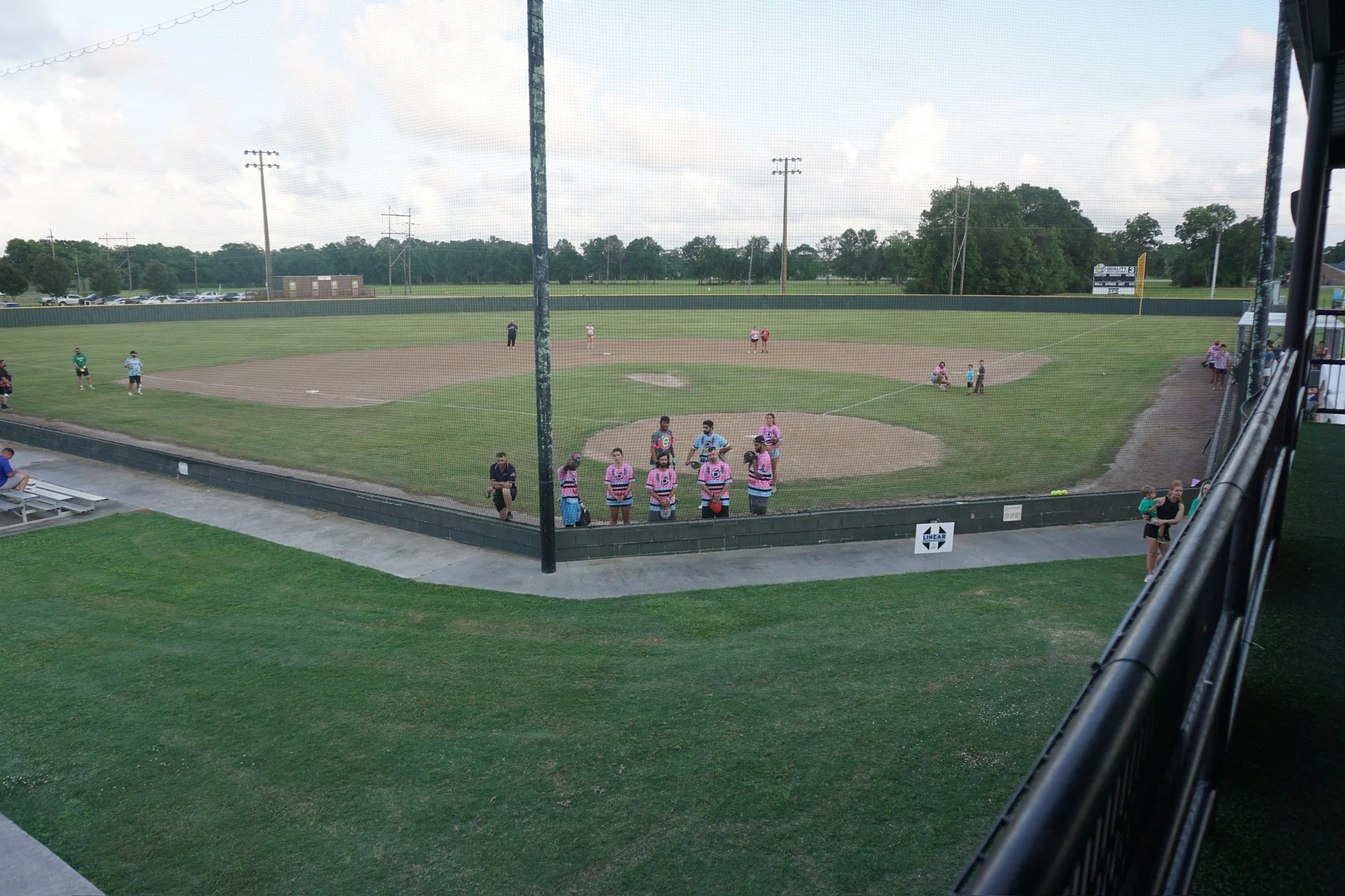 A view of a baseball field from a covered viewing area, with players on the field, spectators on the sidelines, and trees in the background.