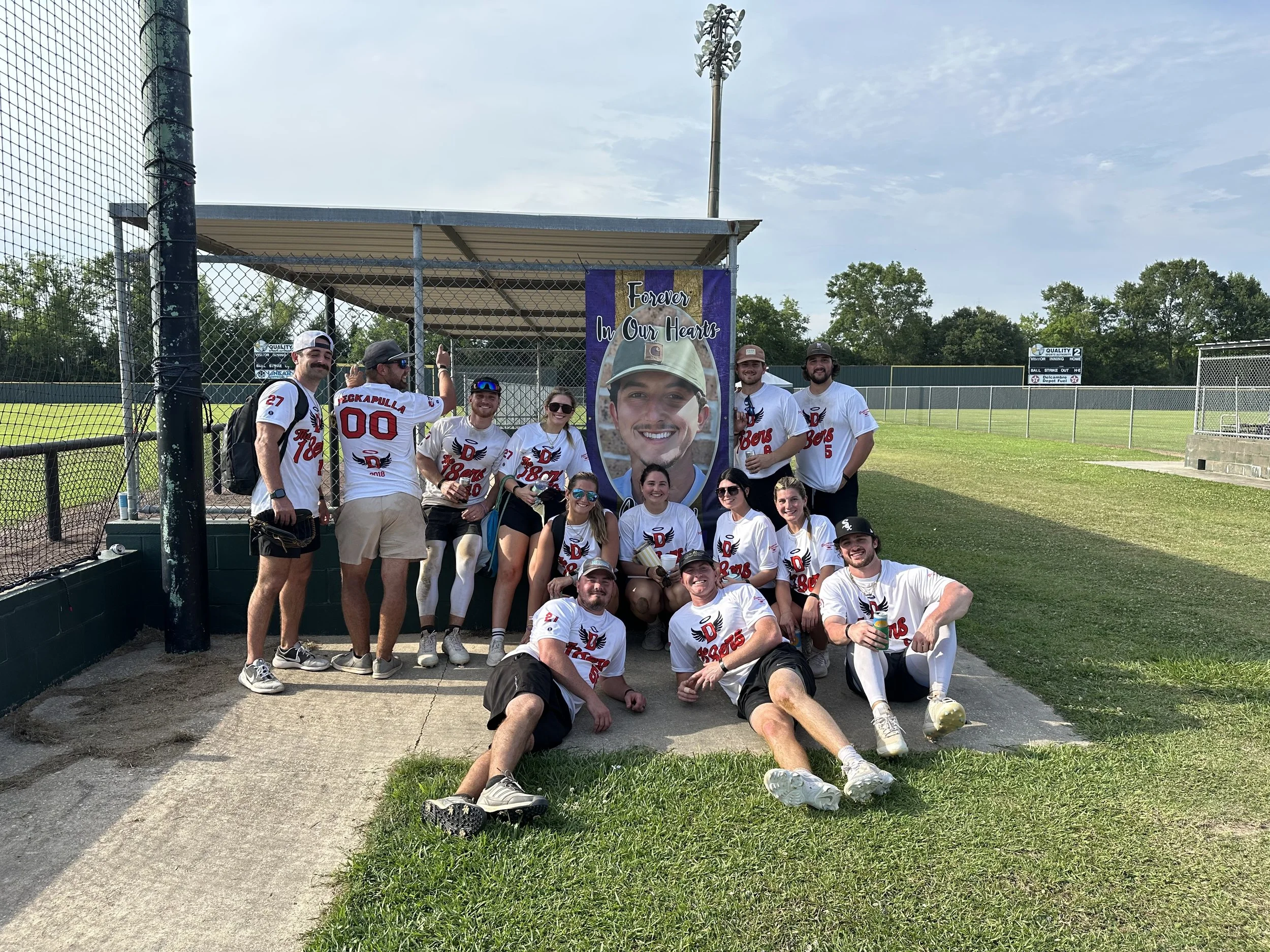 A group of baseball players wearing white jerseys with red and black lettering and numbers, posing together on a baseball field near a large banner with a man's face and the words "Forever in Our Hearts." The team appears happy, some sitting and some