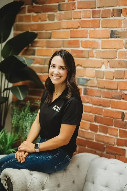A woman sitting on a tufted sofa in front of a brick wall, smiling at the camera, wearing a black polo shirt with a logo, jeans, and a smartwatch, with large green plants in the background.