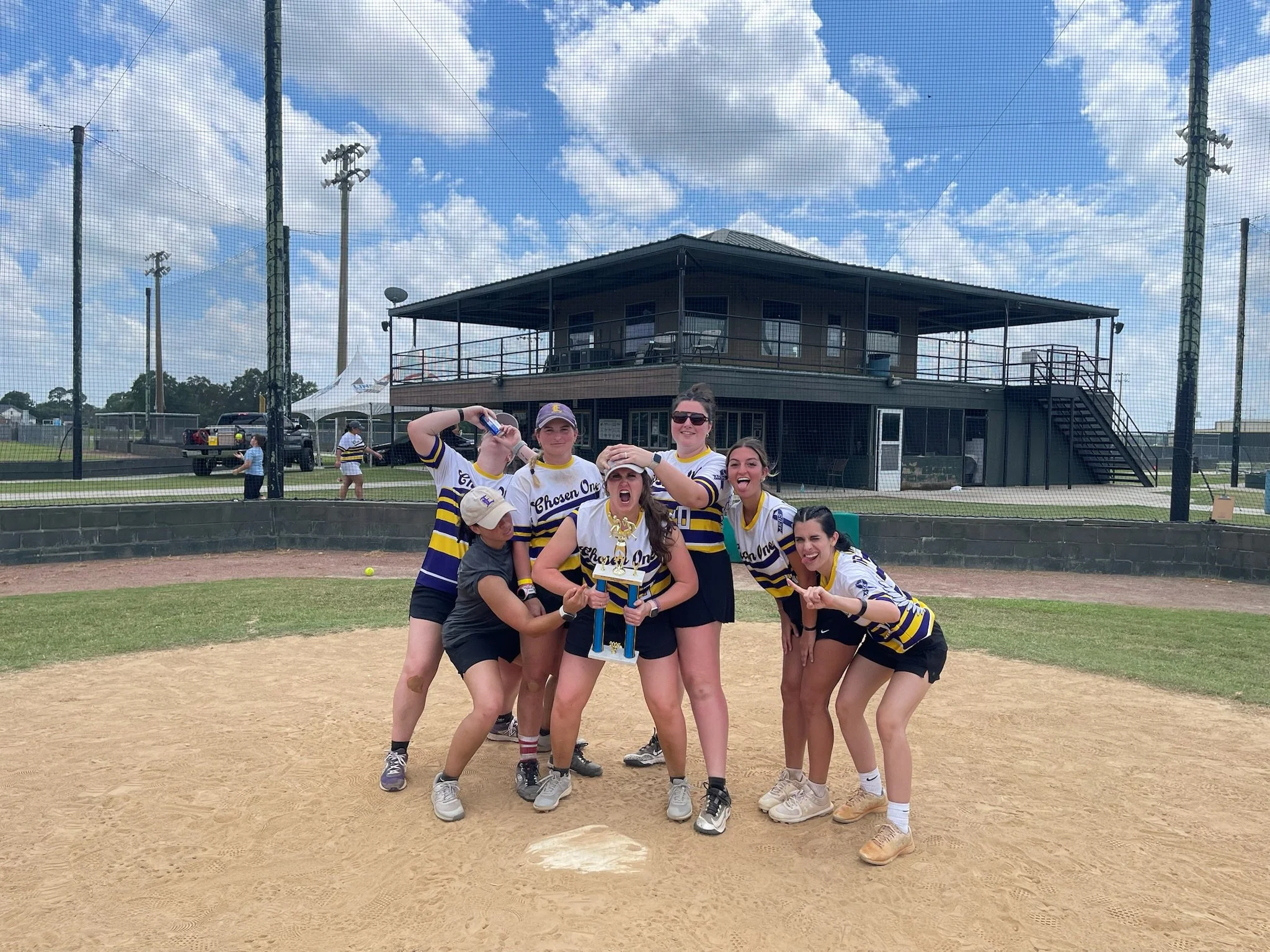 A group of seven girls in baseball uniforms celebrating on a baseball field, holding a trophy and making victory signs, with a building and cloudy sky in the background.