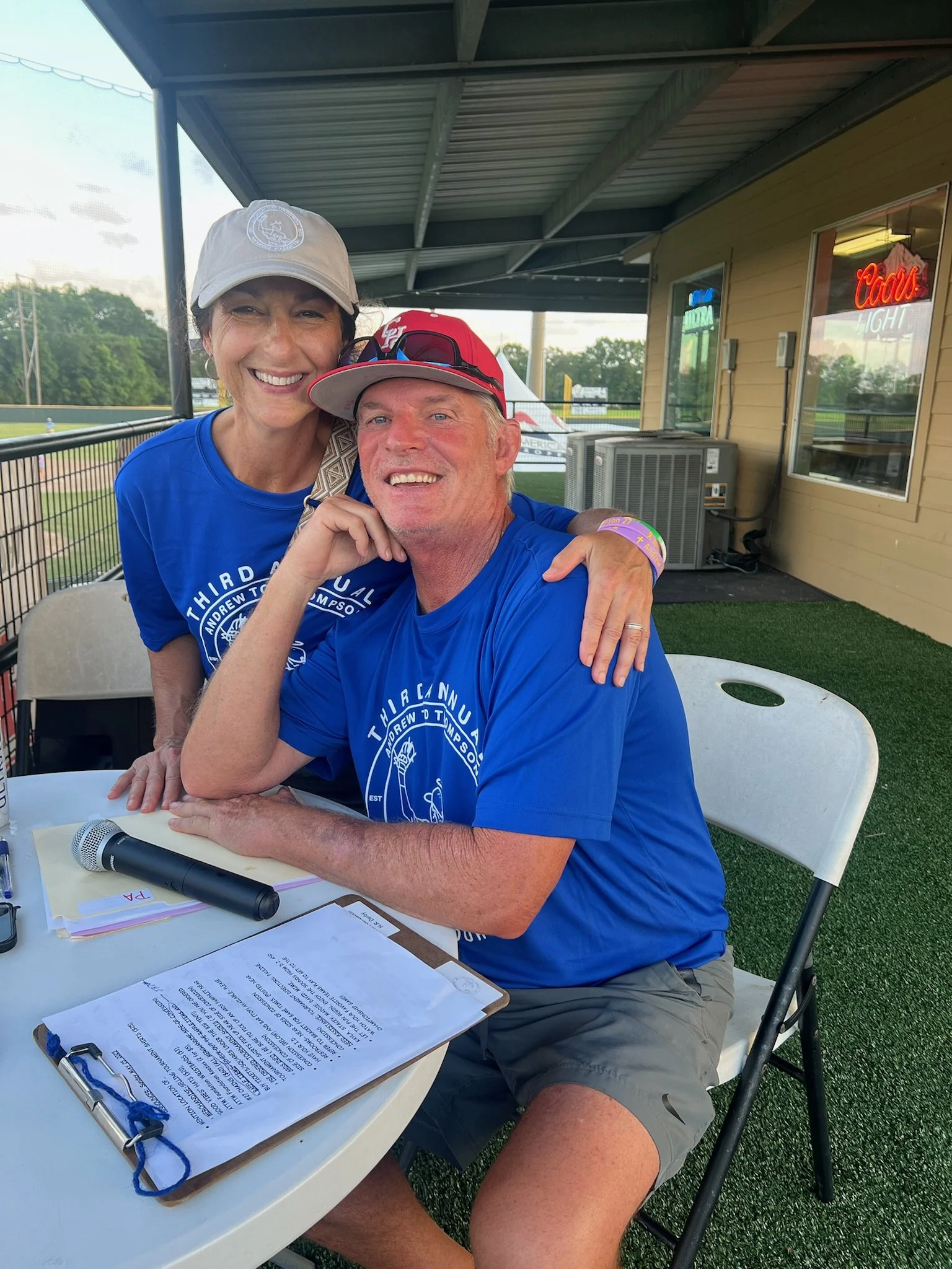 A smiling man and woman wearing blue shirts with a logo, sitting and standing at an outdoor table with a microphone and clipboard, at a baseball stadium.