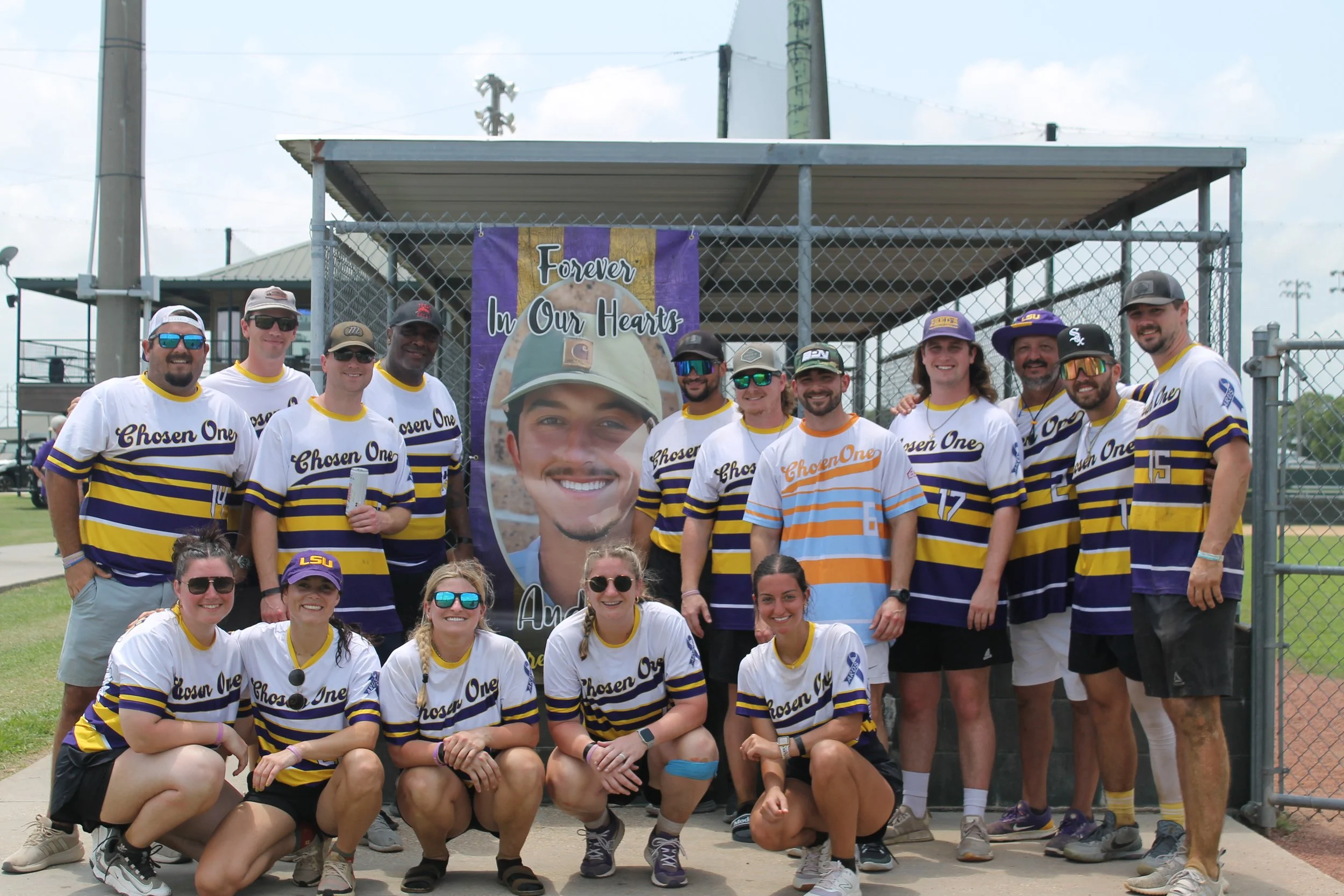 Group of people at a baseball field, wearing matching shirts with 'Chosen One' written on them, standing in front of a memorial banner with a person's photo and the words 'Forever In Our Hearts'.