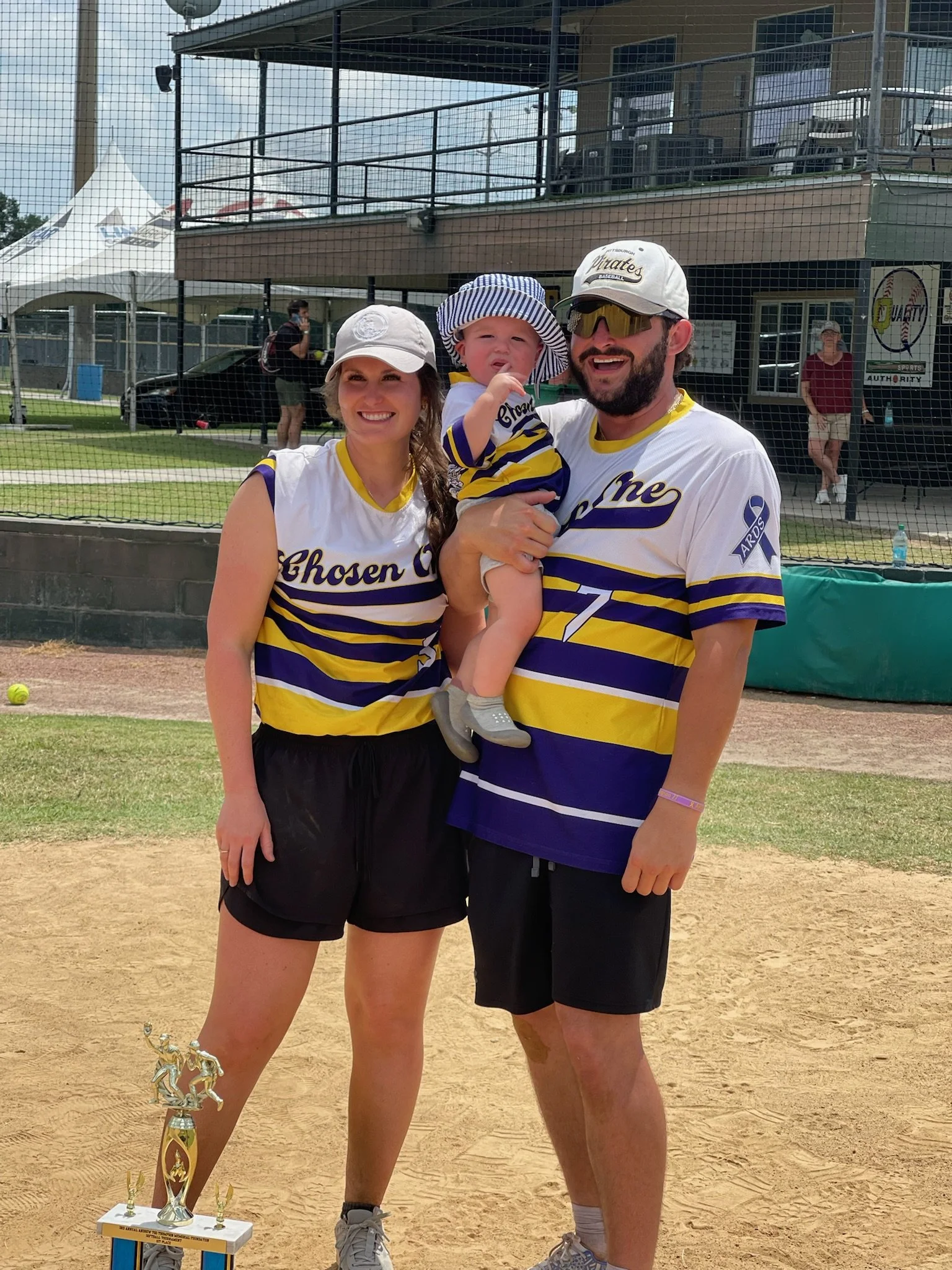 A family of three standing on a baseball field, posing with a trophy. They are wearing matching sports jerseys; the woman and man are holding a young child. The woman is wearing a white cap, the man is wearing sunglasses and a white cap, and the chil