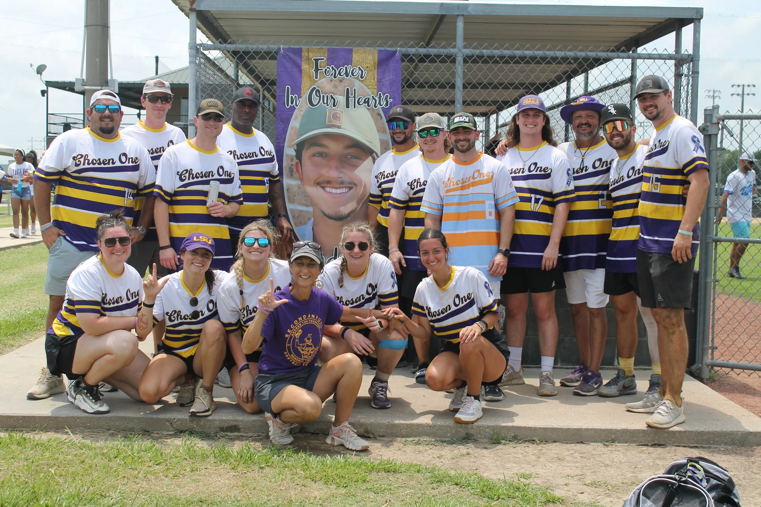 Group of people at a sports event, some wearing LSU gear, gathered in front of a large photo and banner that says 'Forever In Our Hearts,' on a baseball field.