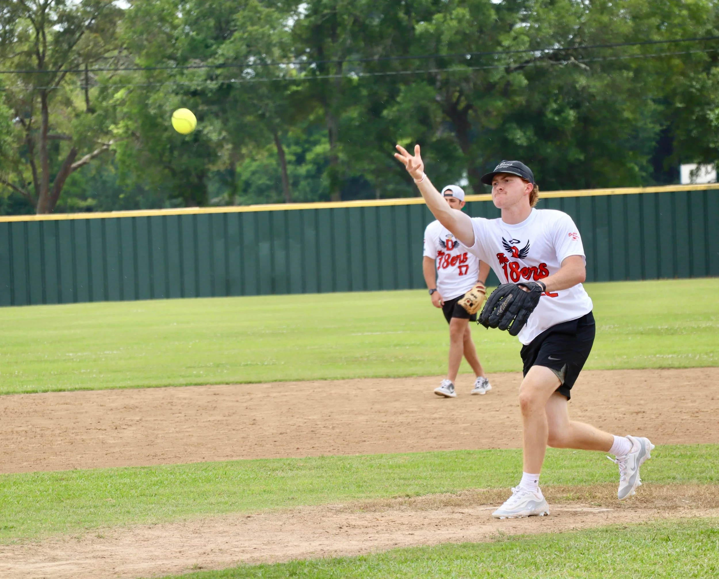 A young man playing baseball on a field, throwing a ball with his left hand while holding a glove with his right hand, wearing a white t-shirt, black shorts, and cap. Another person stands in the background.
