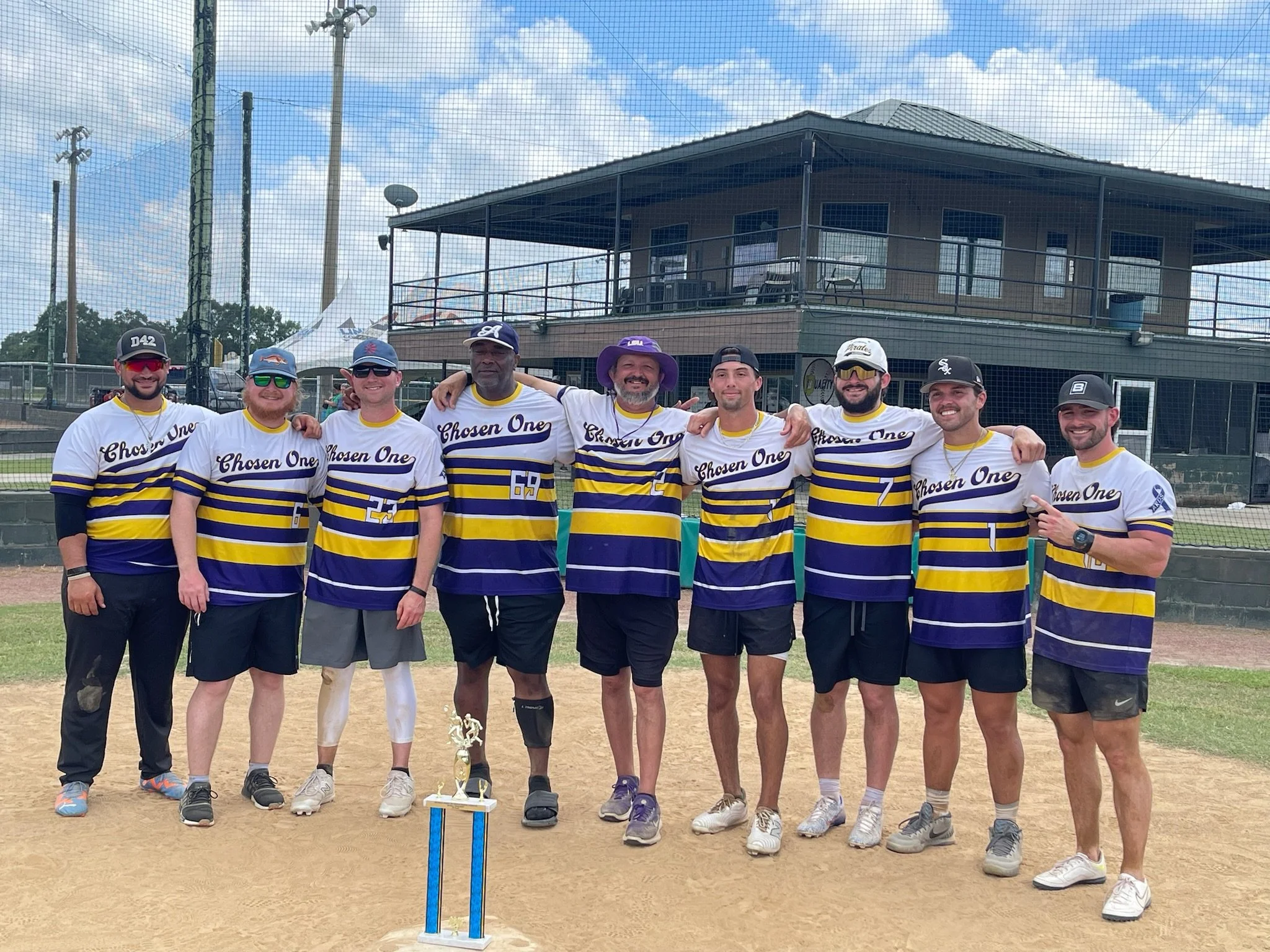 Group of nine men standing on a baseball field, wearing matching jerseys that say 'Chosen One', with a trophy in front of them. They are posing for a team photo, with some men having their arms around each other. In the background, there is a buildin