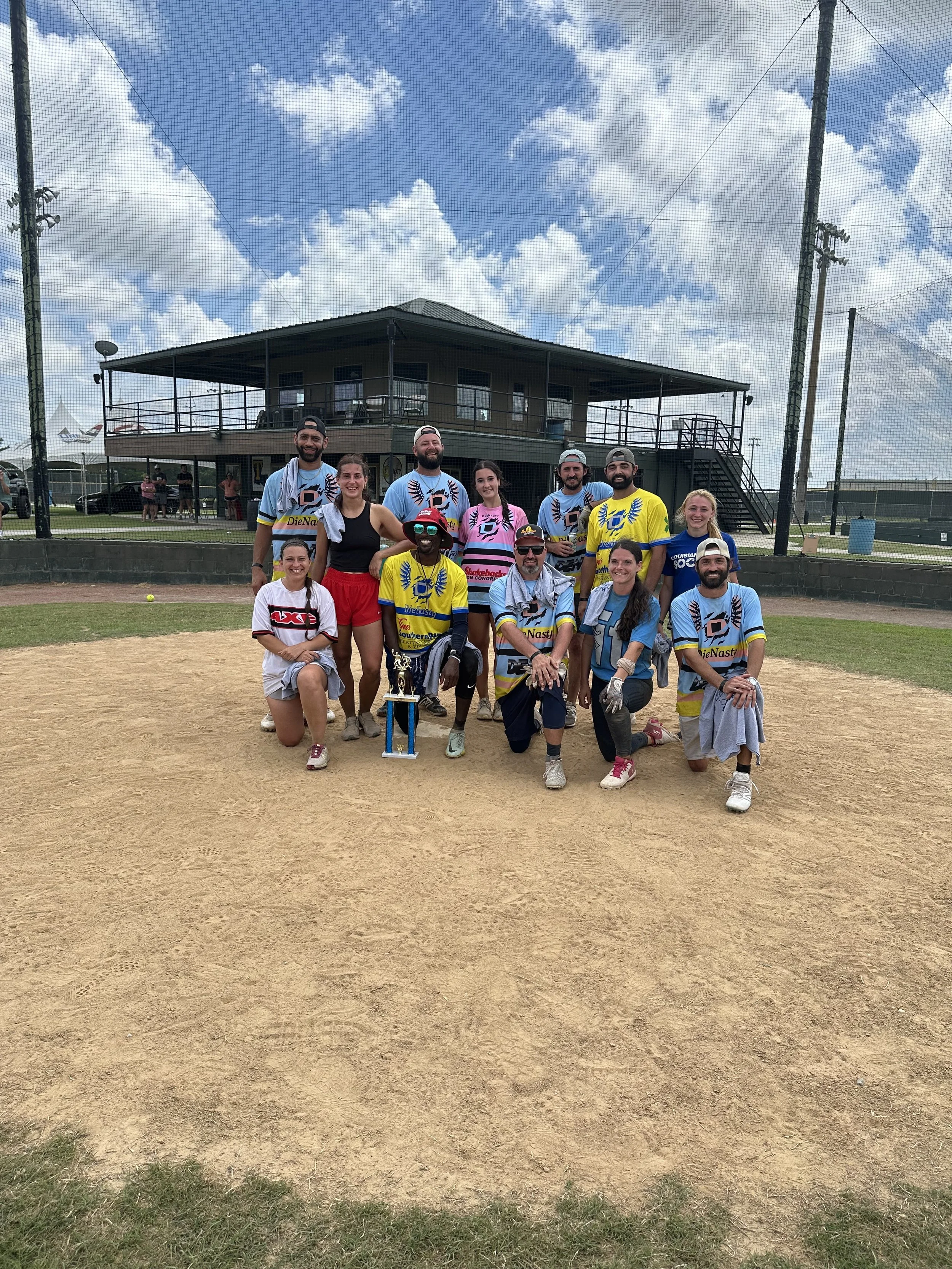 A group of people on a baseball field, some wearing team jerseys, posing for a photo with a trophy. The background shows a clubhouse, baseball net, and a partly cloudy sky.