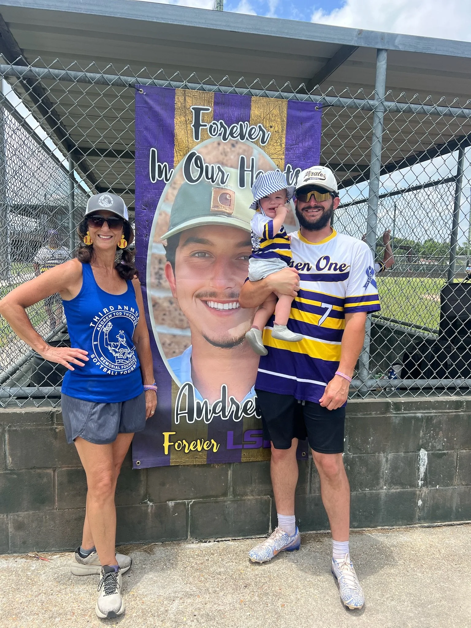 A woman, a man, and a young child stand in front of a purple and gold memorial banner for Andrew, with the words "Forever In Our Hearts" and "Forever LSU" on it. The woman is wearing sunglasses, a gray cap, a blue sleeveless shirt, gray shorts, and r