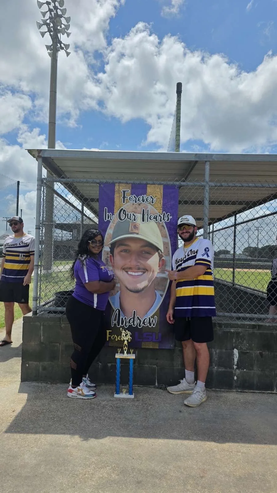 Two people standing in front of a large banner that says 'Forever in Our Hearts Andrew' with a picture of a young man in a military cap. A small trophy is placed on the ground in front of the banner.