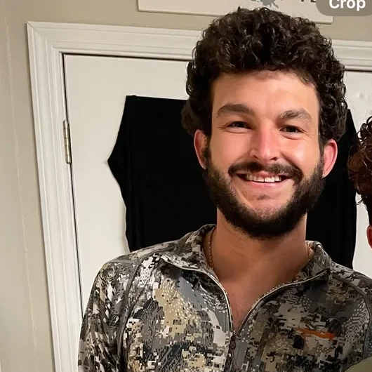 A young man with dark, curly hair and a beard, smiling, wearing a camouflage-patterned zip-up jacket, standing in front of a white door.