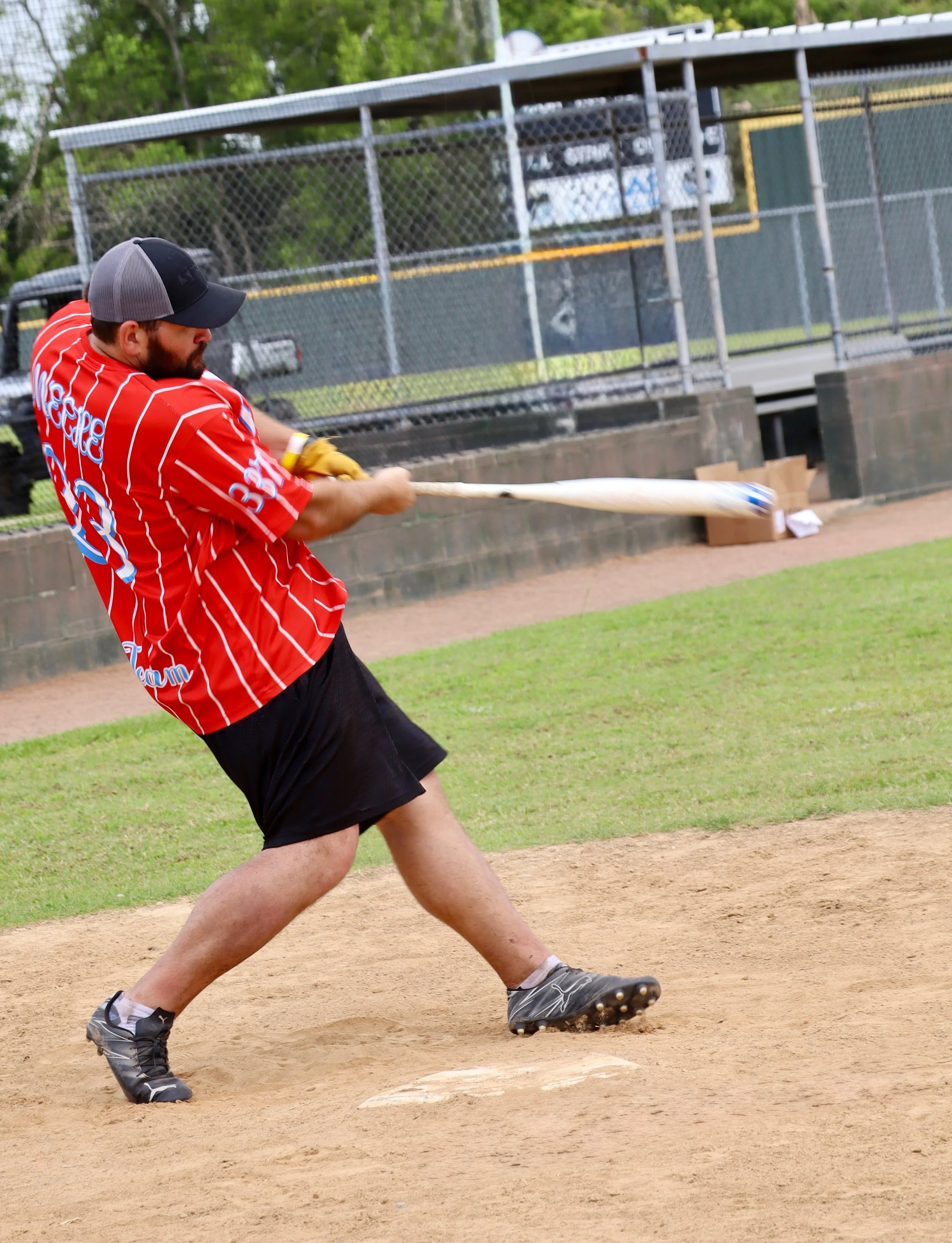 A man batting on a baseball field during a game or practice session, wearing a red and white striped team jersey, black shorts, and a cap.