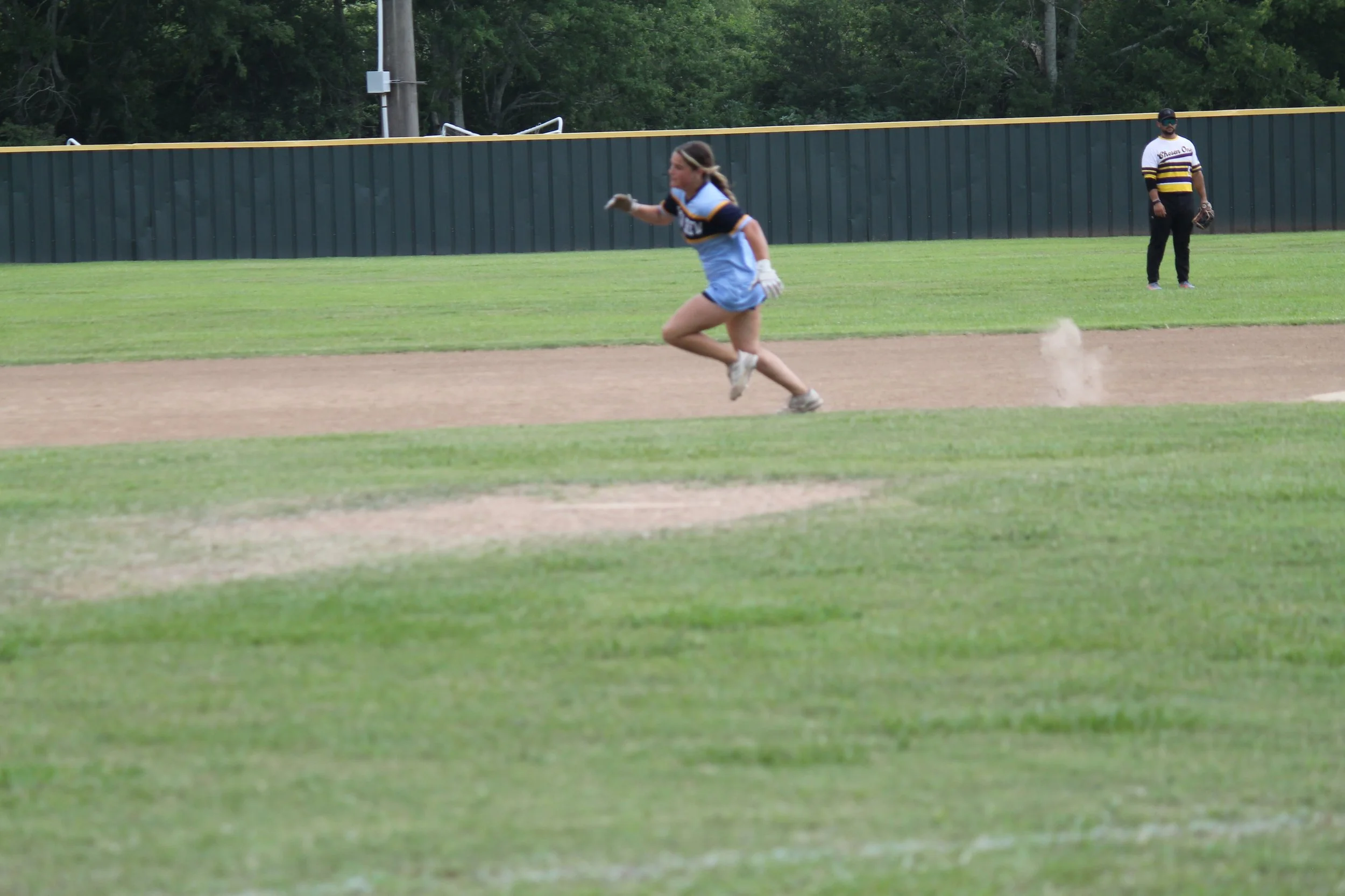 A girl running on a baseball field near third base, with a man standing in the background near the outfield fence, during daytime.