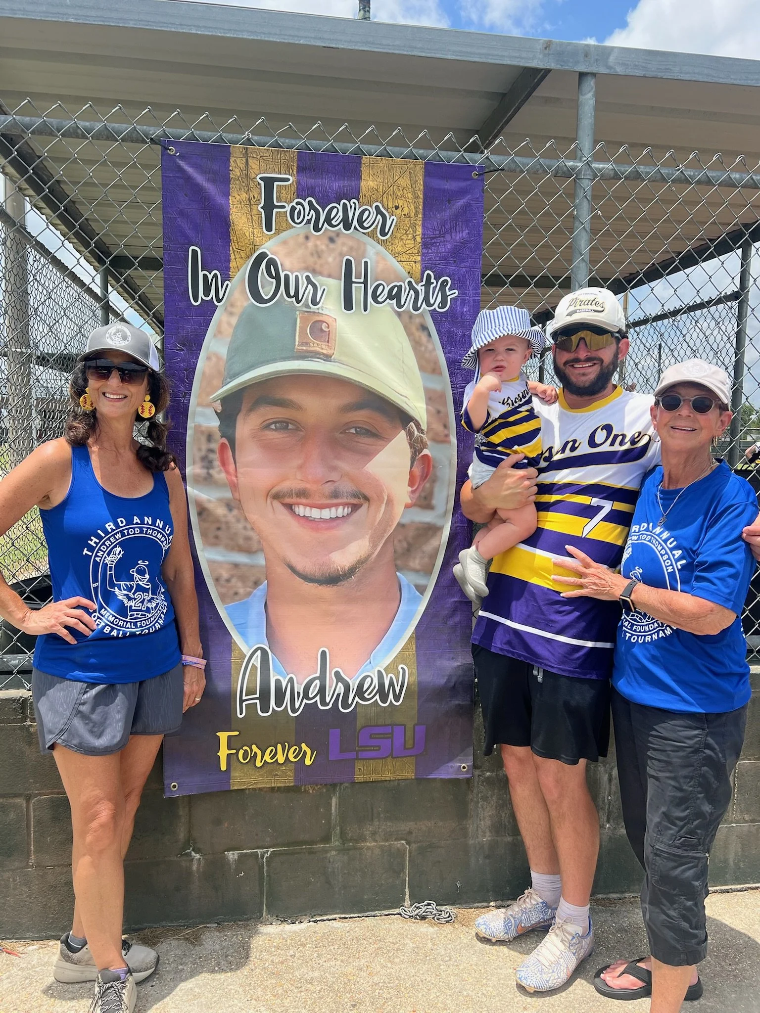 Four people standing outdoors near a chain-link fence with a memorial banner. The banner features a large photo of a young man with the text 'Forever In Our Hearts' and 'Andrew Forever LSU.' The group includes two women, one man, and a young child, a