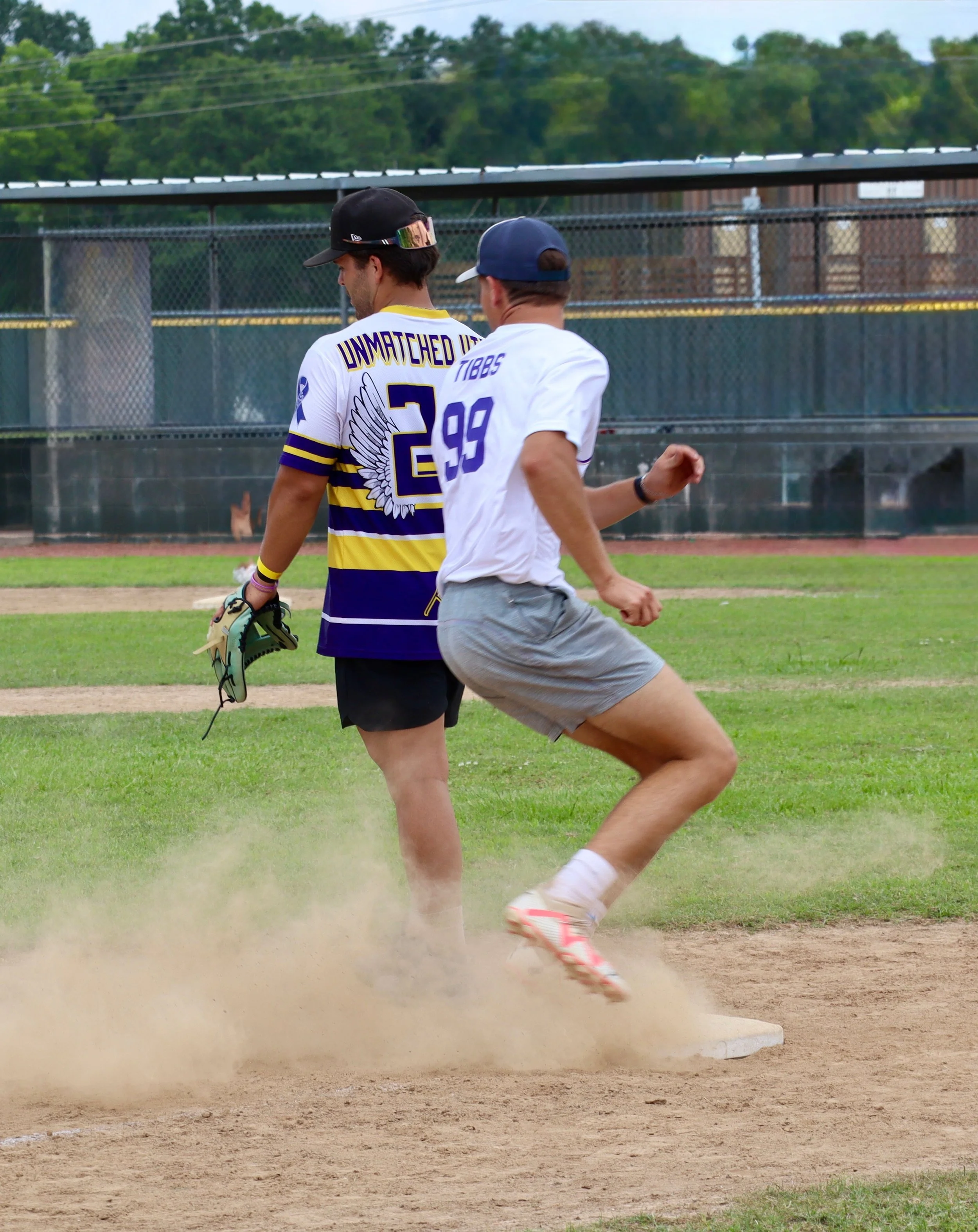 Two young men playing baseball on a field, one is standing at a base with a glove, the other is running past the base.