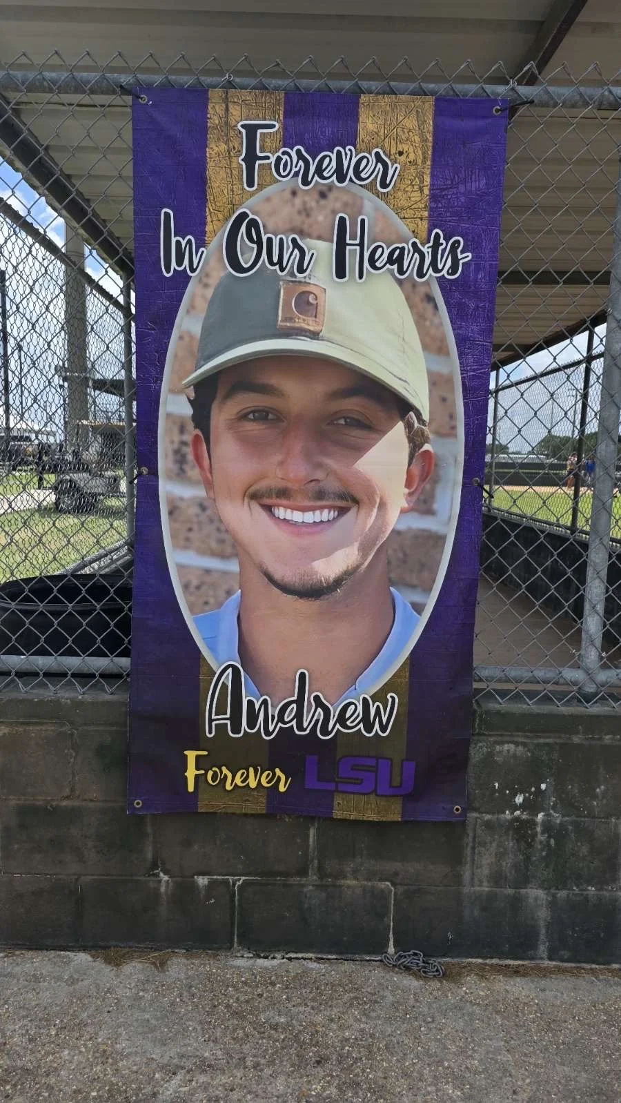 A tribute banner showing a smiling young man with a beard and mustache, wearing a beige cap with the LSU logo, and text that reads 'Forever In Our Hearts Andrew Forever LSU.' The banner has purple, yellow, and gold colors and is attached to a chain-l