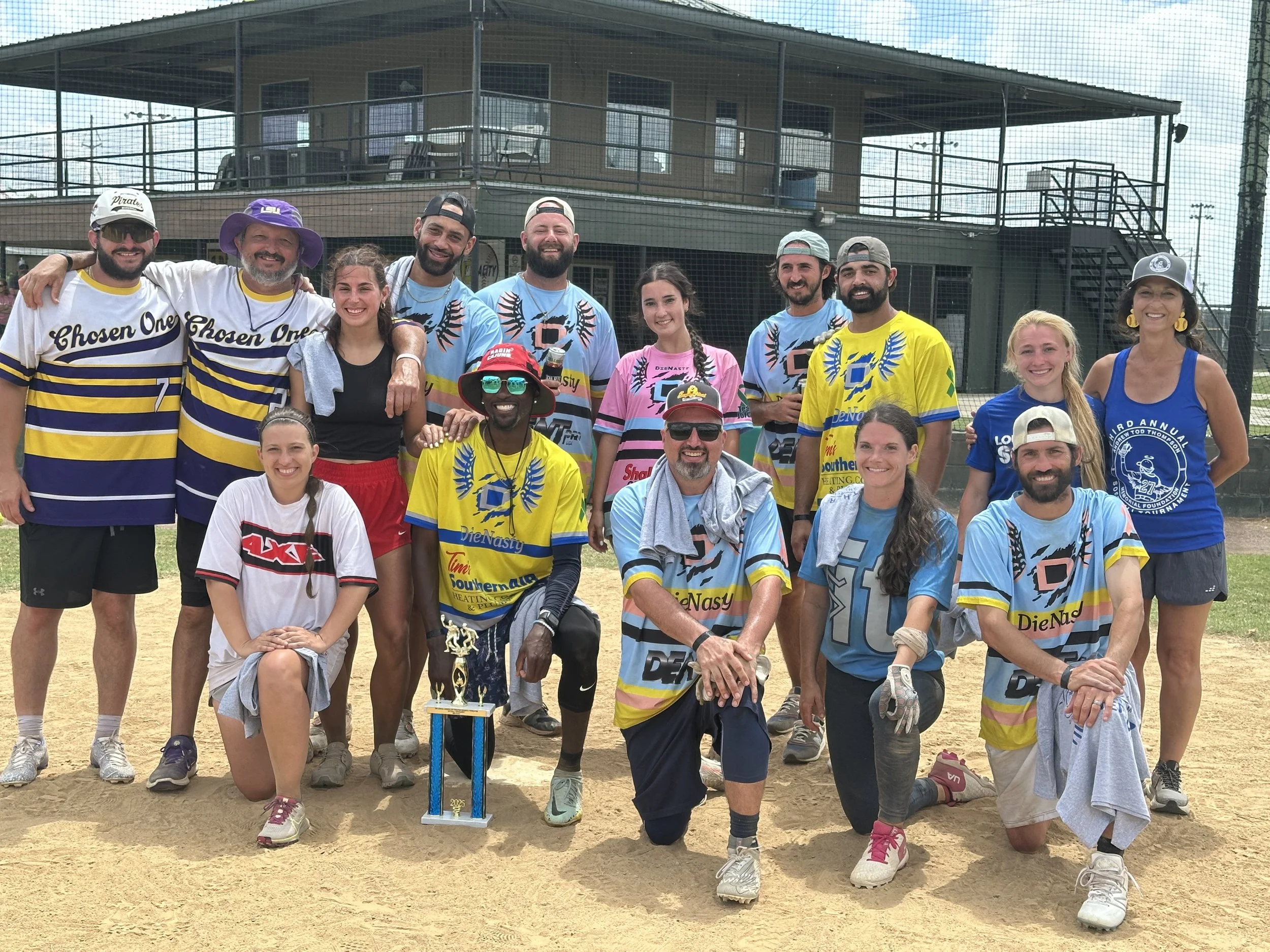 Group of people on a baseball field posing with a trophy, wearing colorful sports jerseys and casual clothing, with a clubhouse in the background.