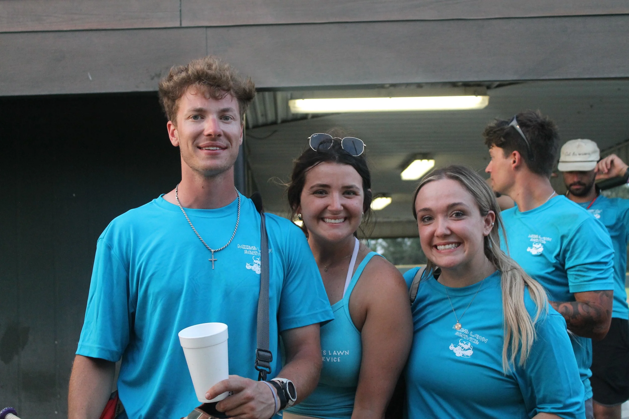 Group of four young adults smiling outdoors, wearing matching blue T-shirts, with two women in the center and two men on the sides, in a casual setting.